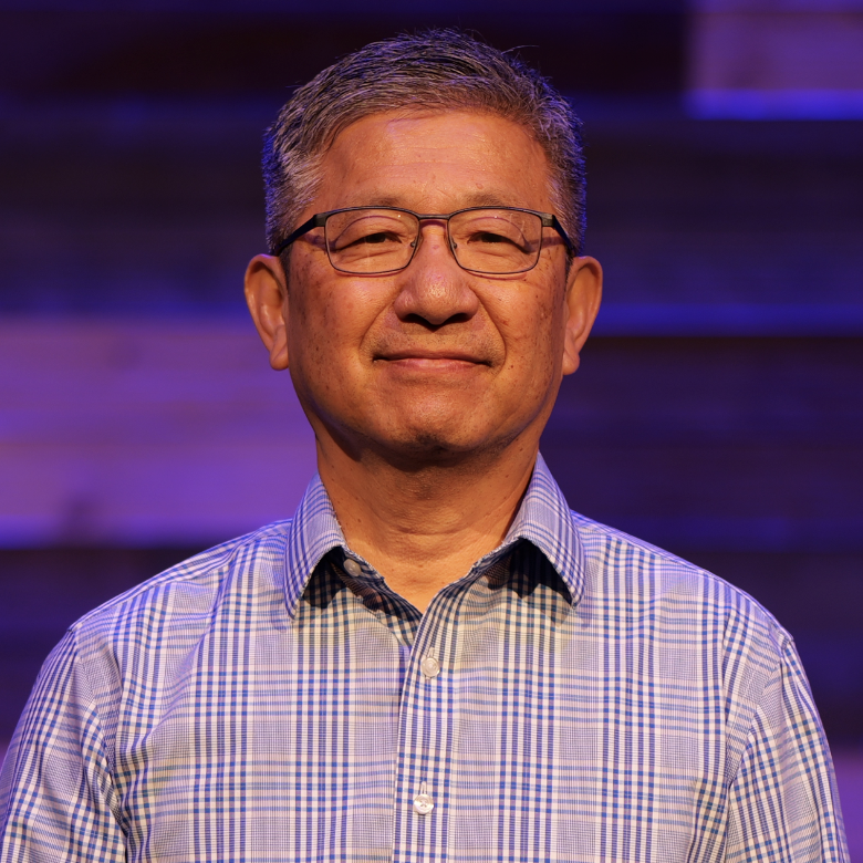 A middle-aged Asian man with glasses and short gray hair, smiling, wearing a blue checkered shirt, standing against a purple and blue blurred background.