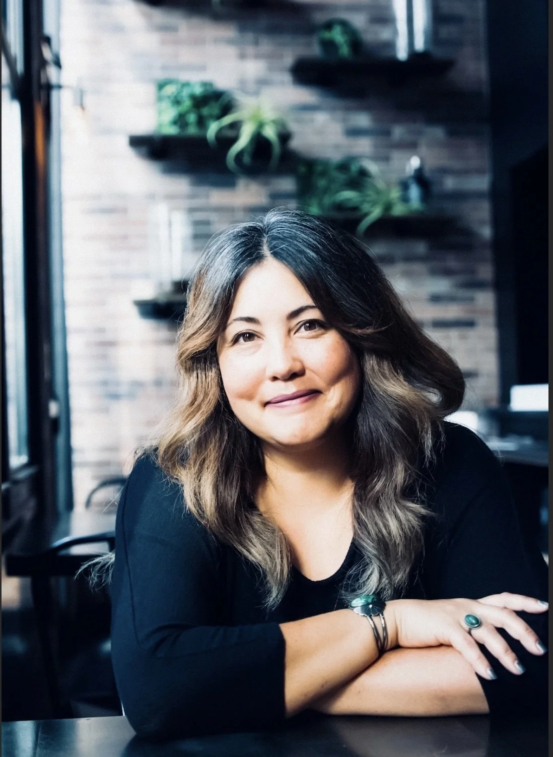 A woman with wavy, shoulder-length hair, wearing a black top and jewelry, is sitting at a table in a cafe or restaurant, smiling at the camera. The background features a brick wall with shelves and plants.
