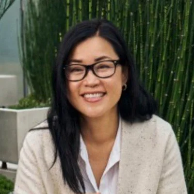 A woman with long black hair, glasses, and a white shirt smiling outdoors surrounded by green plants.