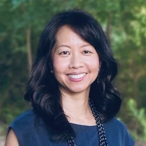 A woman with shoulder-length black hair wearing a navy blue top and beaded necklace standing outdoors with green trees in the background.