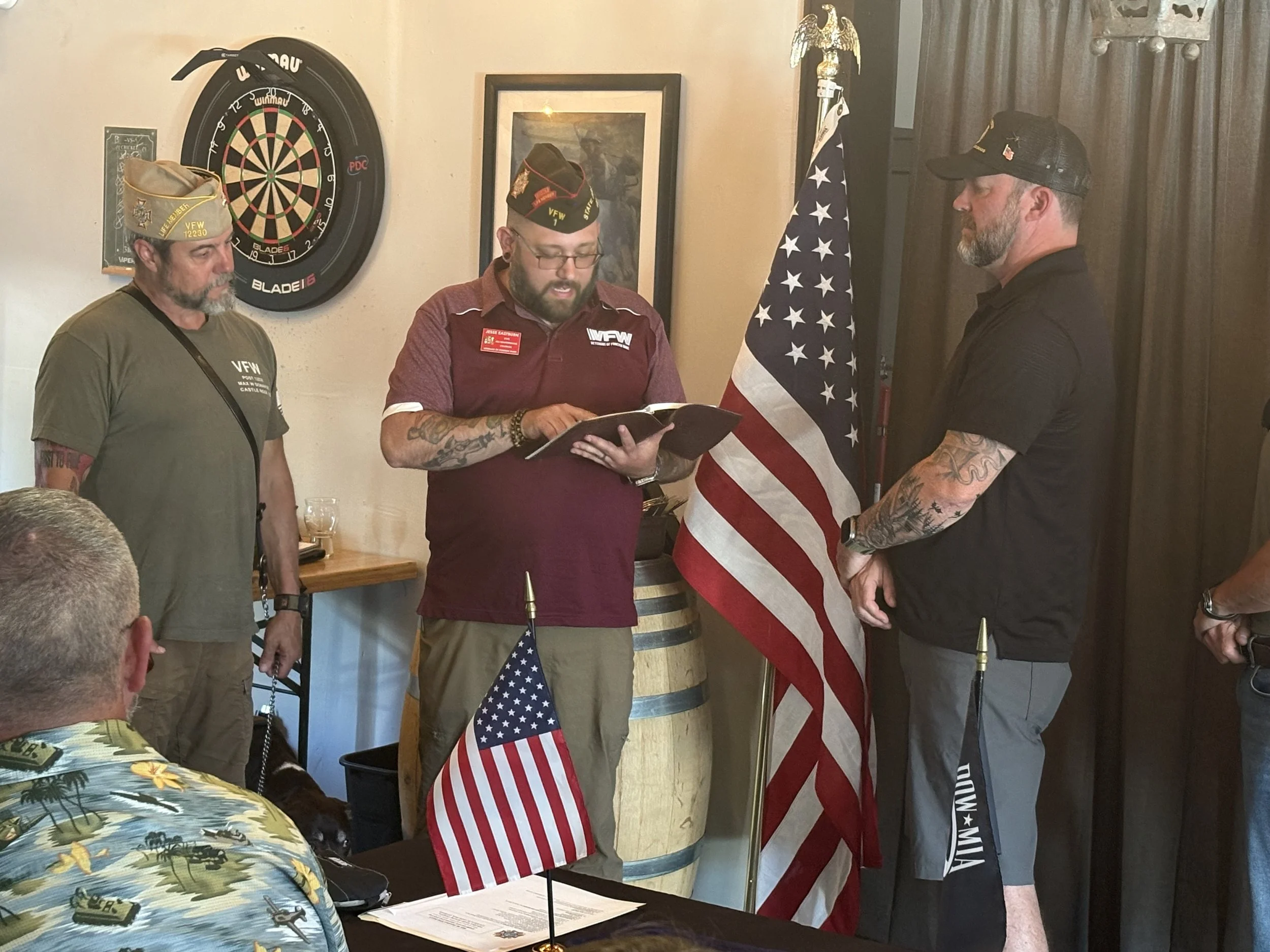 Three men wearing Veterans of Foreign Wars (VFW) hats stand in front of an American flag, participating in a ceremony or oath-taking, with a dartboard on the wall and a person seated in the foreground.