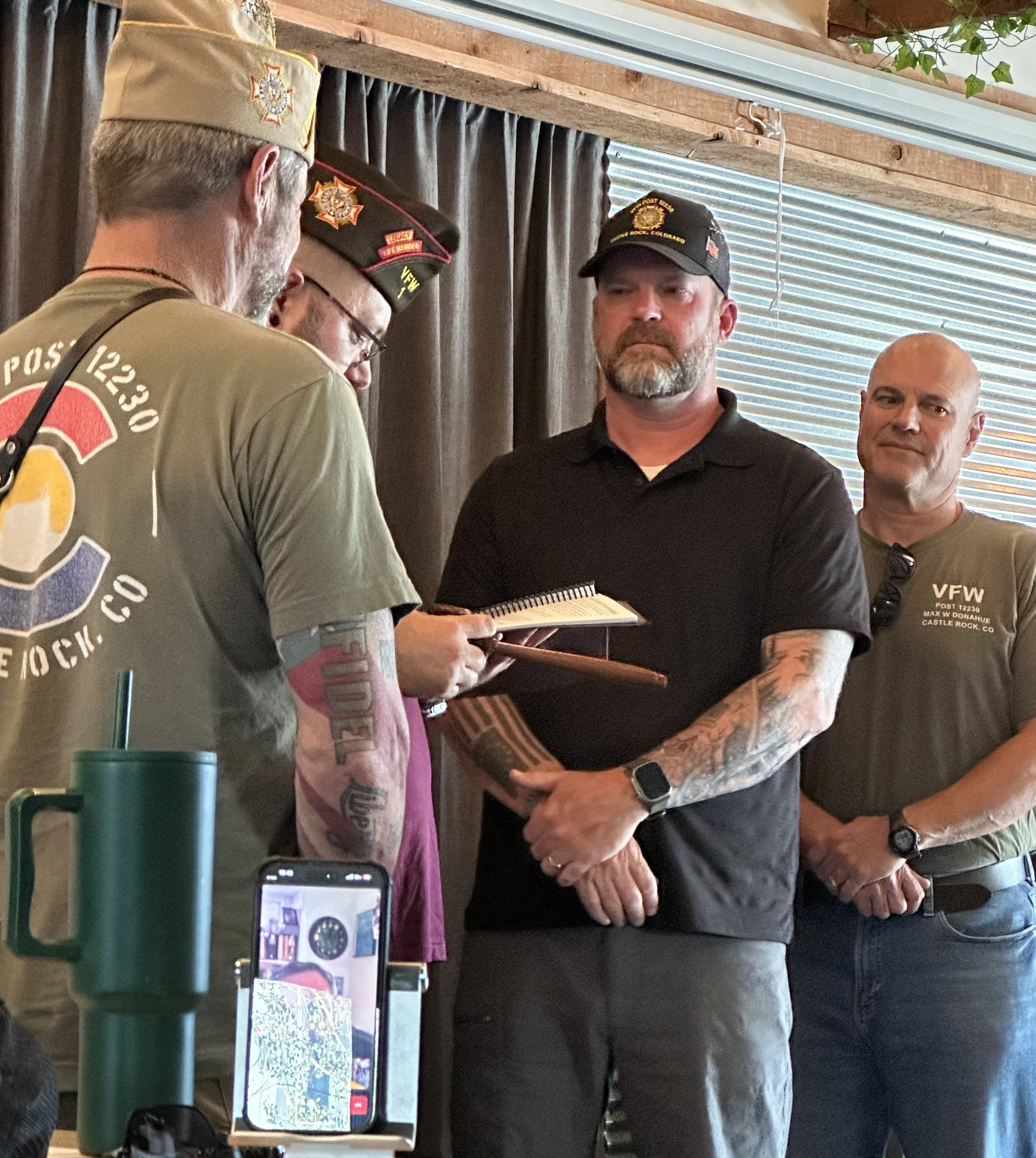 Three men standing indoors, with two wearing American Legion hats, the man in the center wearing a black shirt, and one man wearing a gray T-shirt with 'VFW' and 'Post 1220' printed on it. The man on the left is writing in a small booklet, while the 