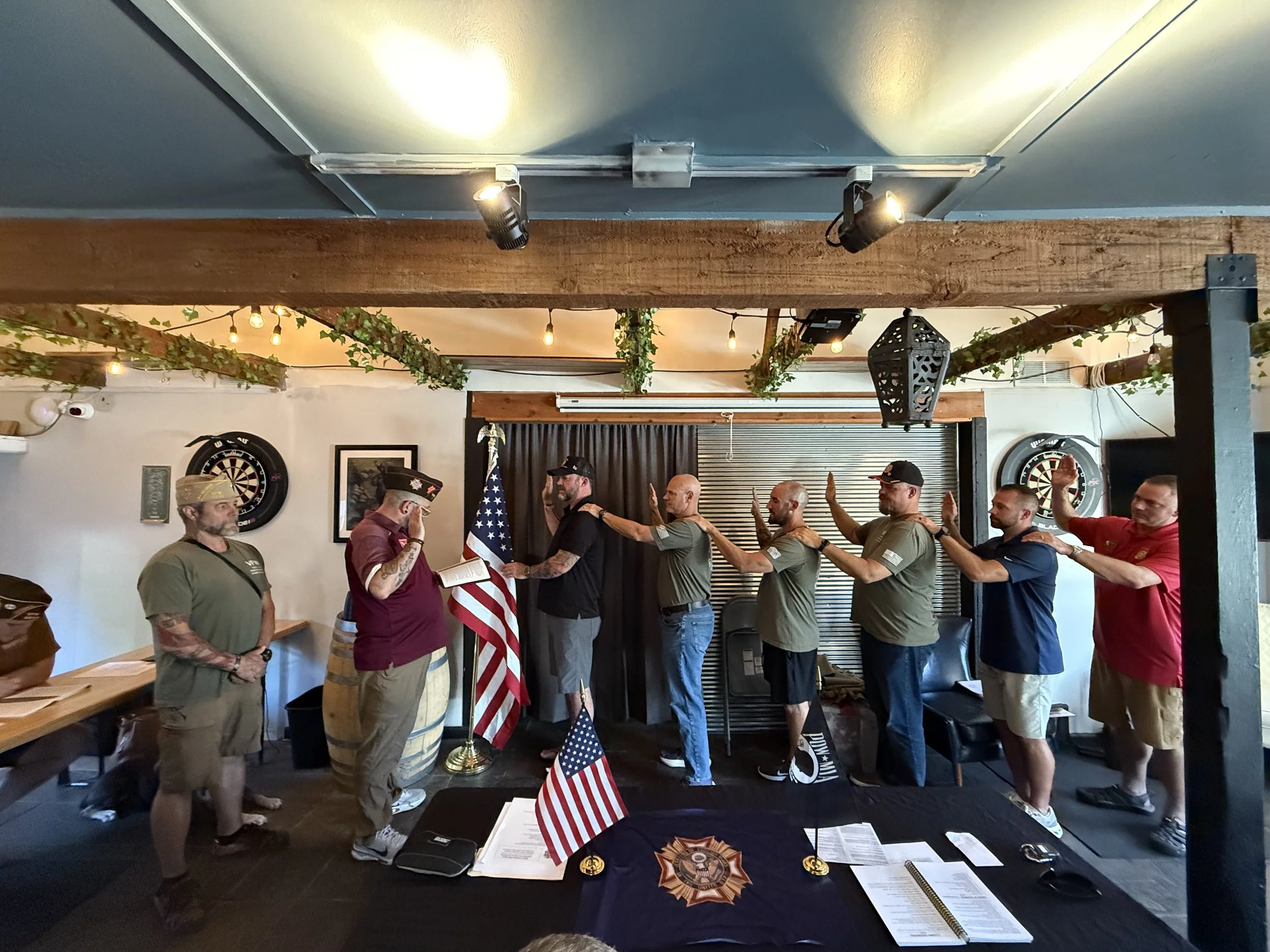 A group of men participating in an oath-taking ceremony inside a room decorated with American flags, dartboards, and string lights, with a table displaying documents and small flags in the foreground.