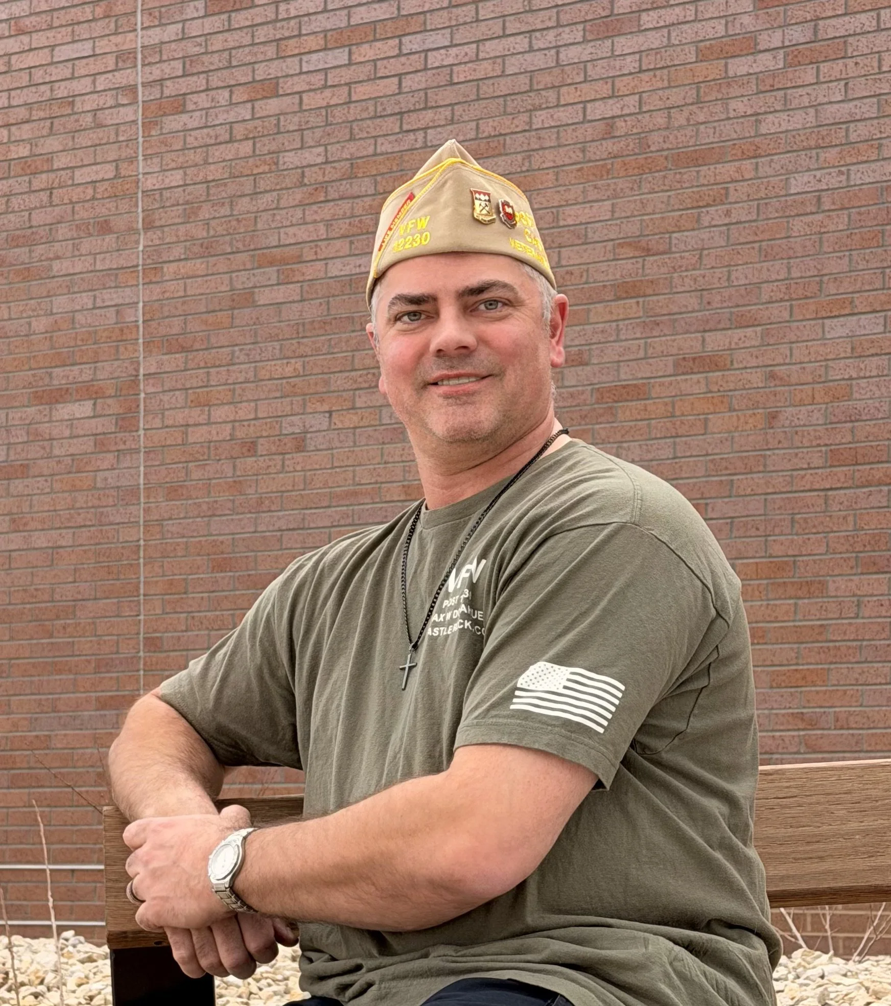 A man sitting on a wooden bench in front of a brick wall, wearing a beige military cap, a gray T-shirt with American flag patch on the sleeve, a wristwatch, and a necklace with a cross pendant.