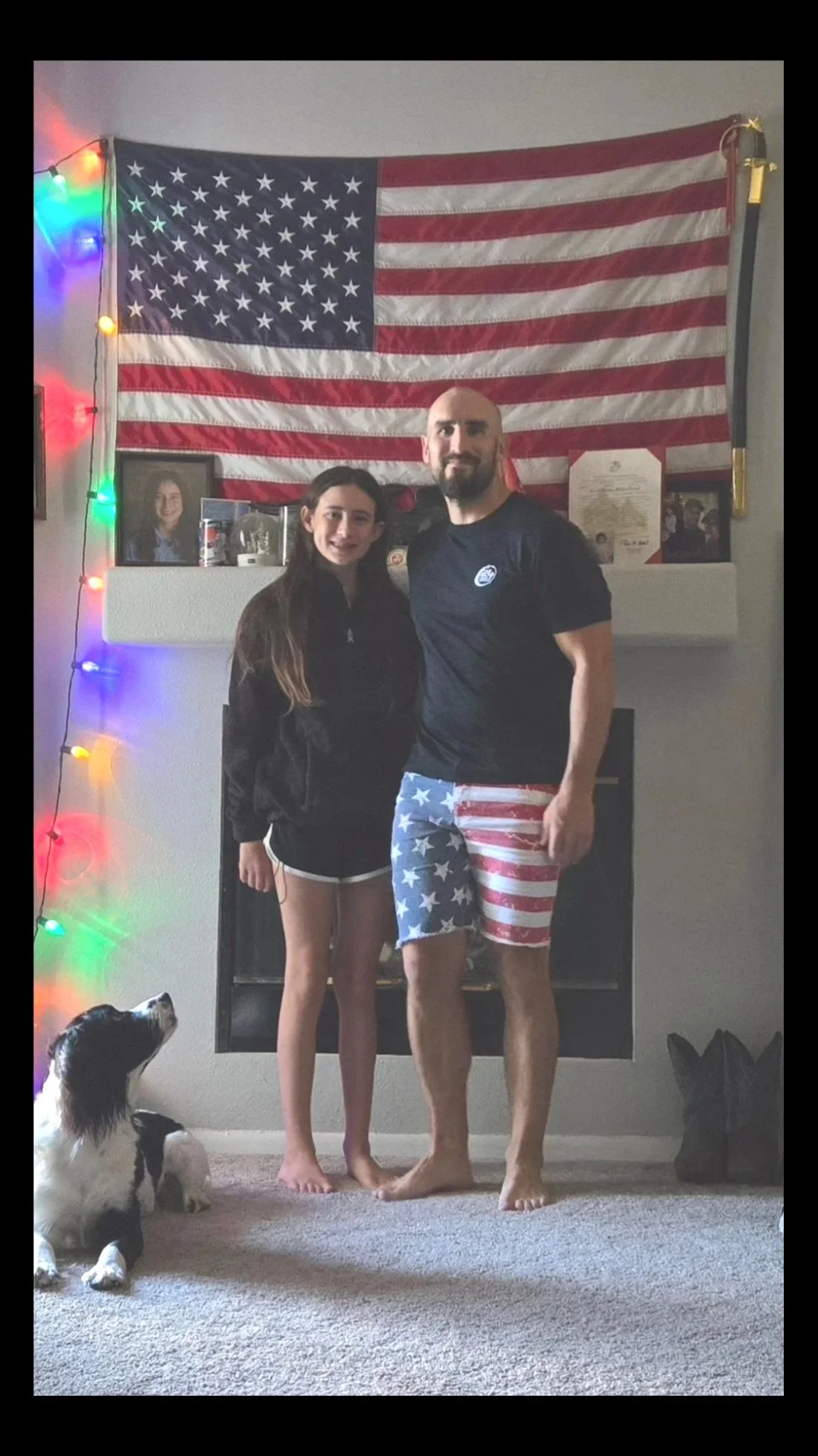 A young girl and an adult man standing barefoot in a living room in front of an American flag hanging on the wall. The man is wearing American flag-themed shorts and a black T-shirt, while the girl is dressed in black shorts and a black jacket. To their left is a dog looking up. There are colorful string lights hanging on the wall and framed photos and certificates on a shelf behind them.