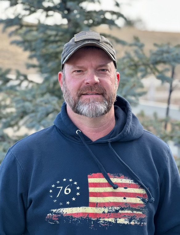Man with a beard wearing a navy hoodie with a distressed American flag and the number 76, standing outdoors with trees in the background.