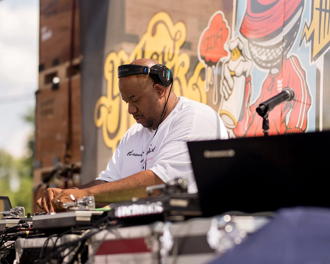 A DJ wearing headphones is performing outdoors, with a colorful mural featuring a cartoon character in the background and a microphone on a stand nearby.