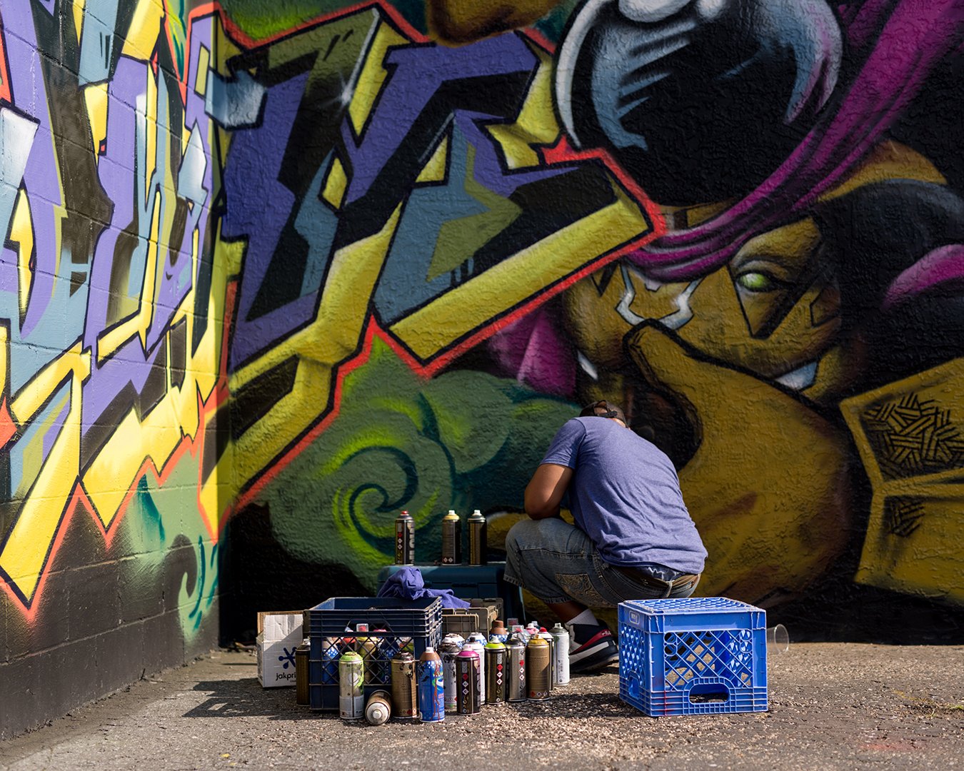 A person crouched down spray painting graffiti on a large wall that features a colorful mural of a face with purple hair and a green hand. There are spray paint cans and crates on the ground around the artist.