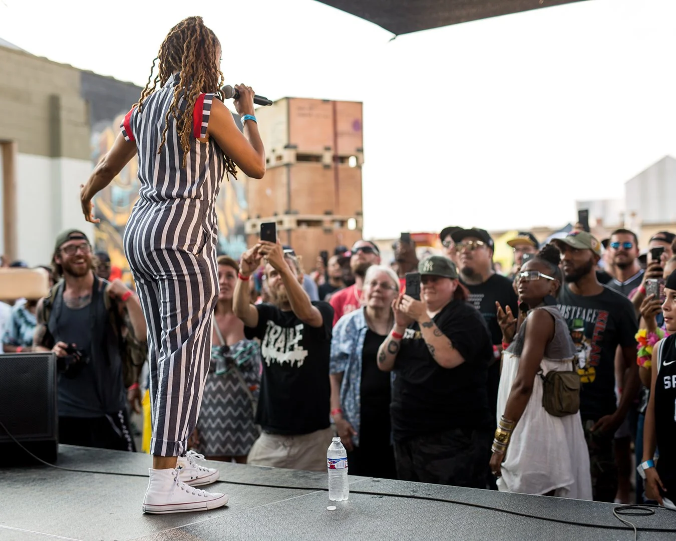 A woman with long dreadlocks, wearing a striped sleeveless jumpsuit and white sneakers, is performing on stage with a microphone in front of an audience at an outdoor event. The crowd is taking photos and videos, some smiling and cheering.