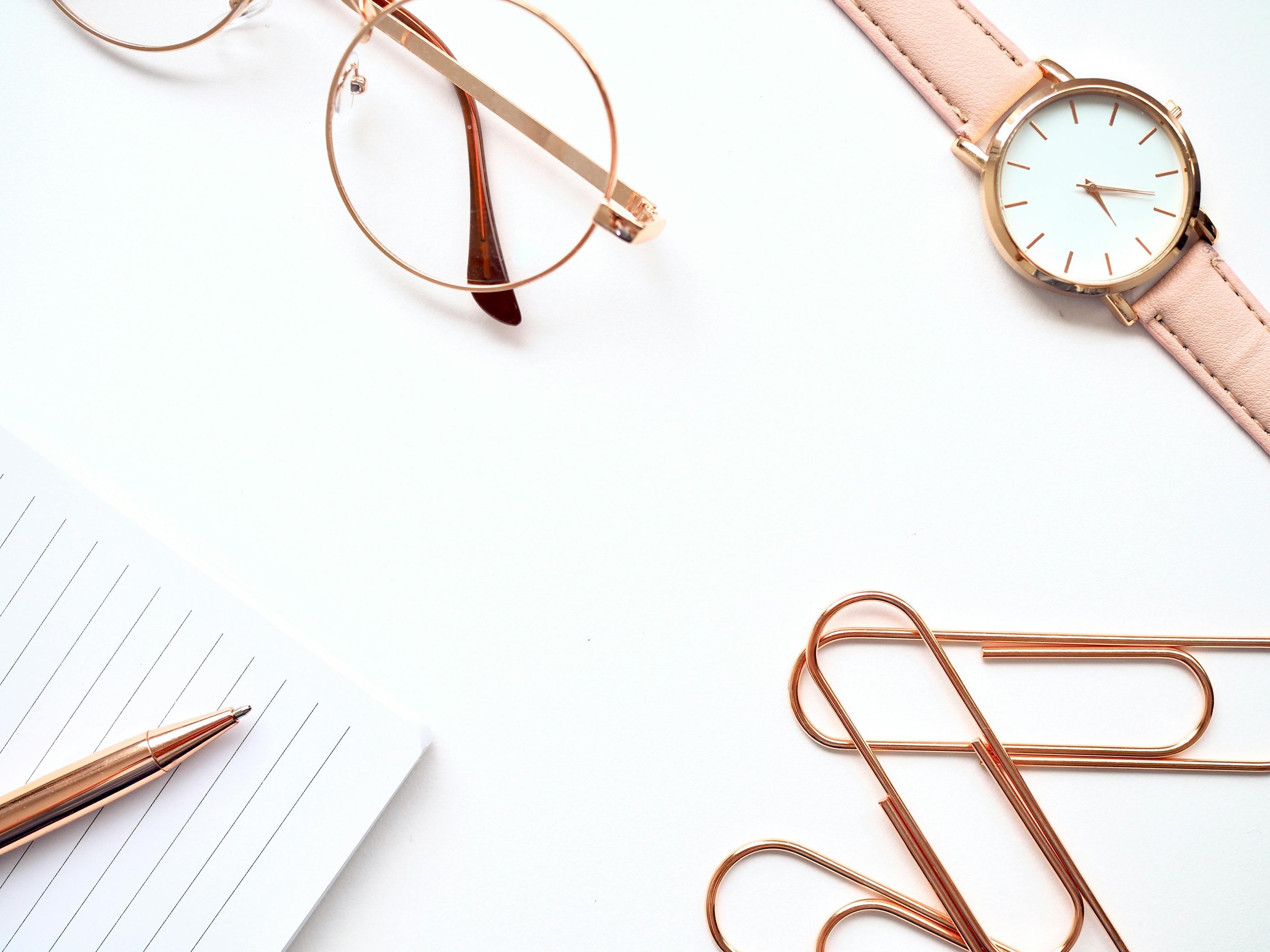 A flat lay of a minimalist workspace featuring a white notebook with black lines and a gold pen, rose gold glasses, a rose gold wristwatch with a pink leather strap, and rose gold paperclips on a white background.