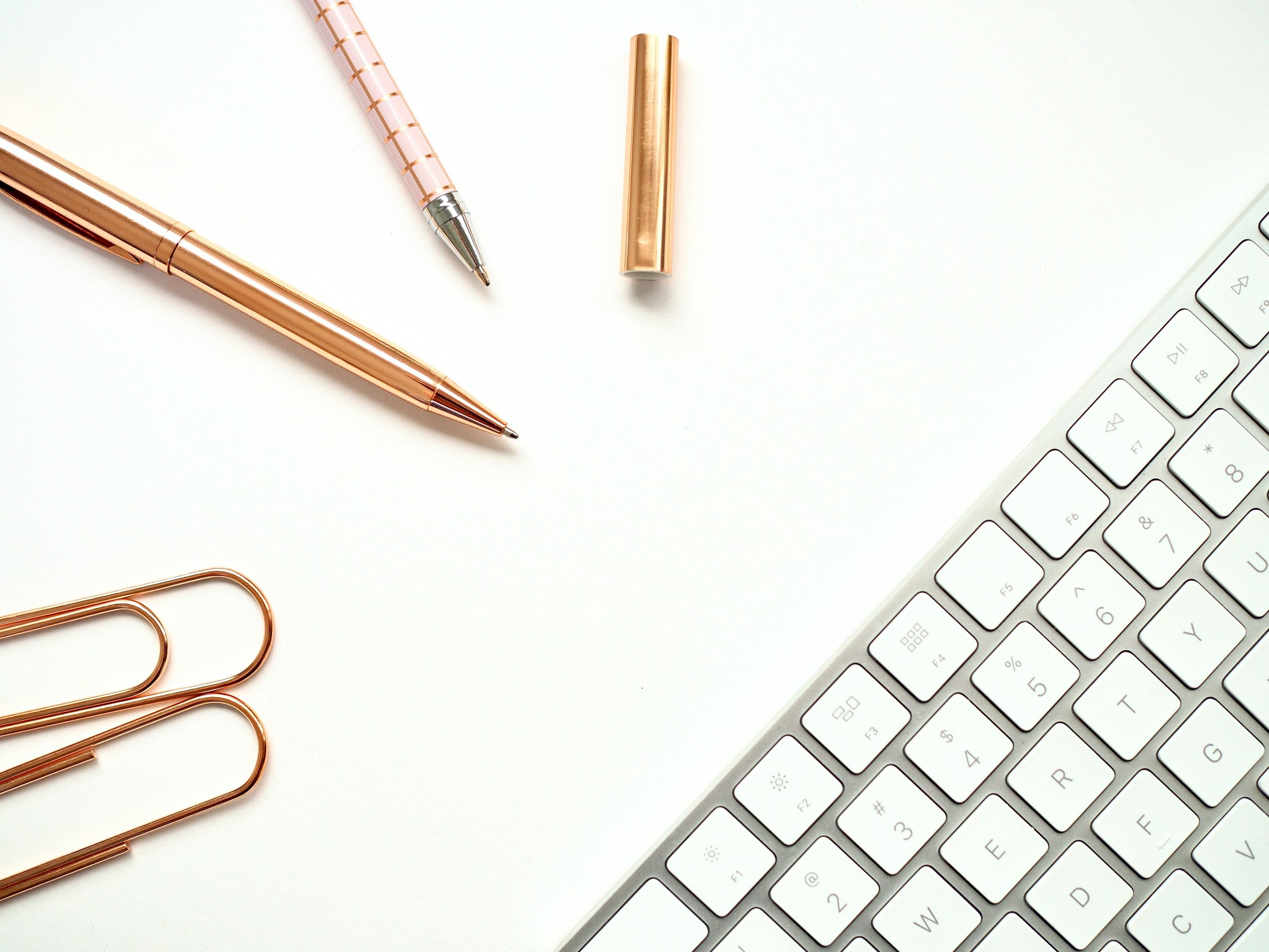 A flat lay of gold and pink pens, paper clips, a pen cap, and a white computer keyboard on a white surface.