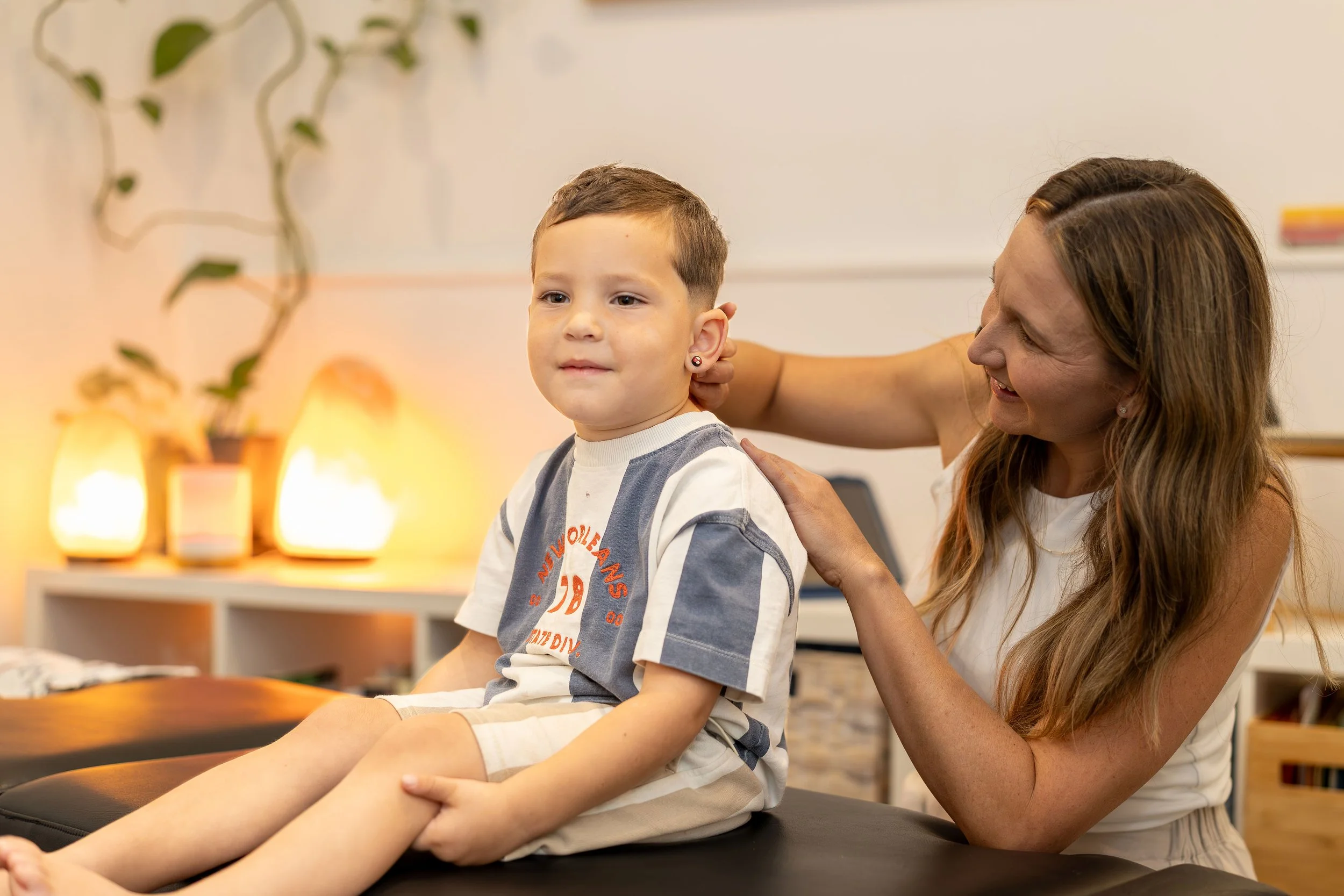 A woman with long reddish-brown hair sitting beside a young boy with short brown hair, as she holds his shoulder and neck, possibly performing a physical therapy or chiropractic adjustment in a room with warm lighting and decorative lamps.