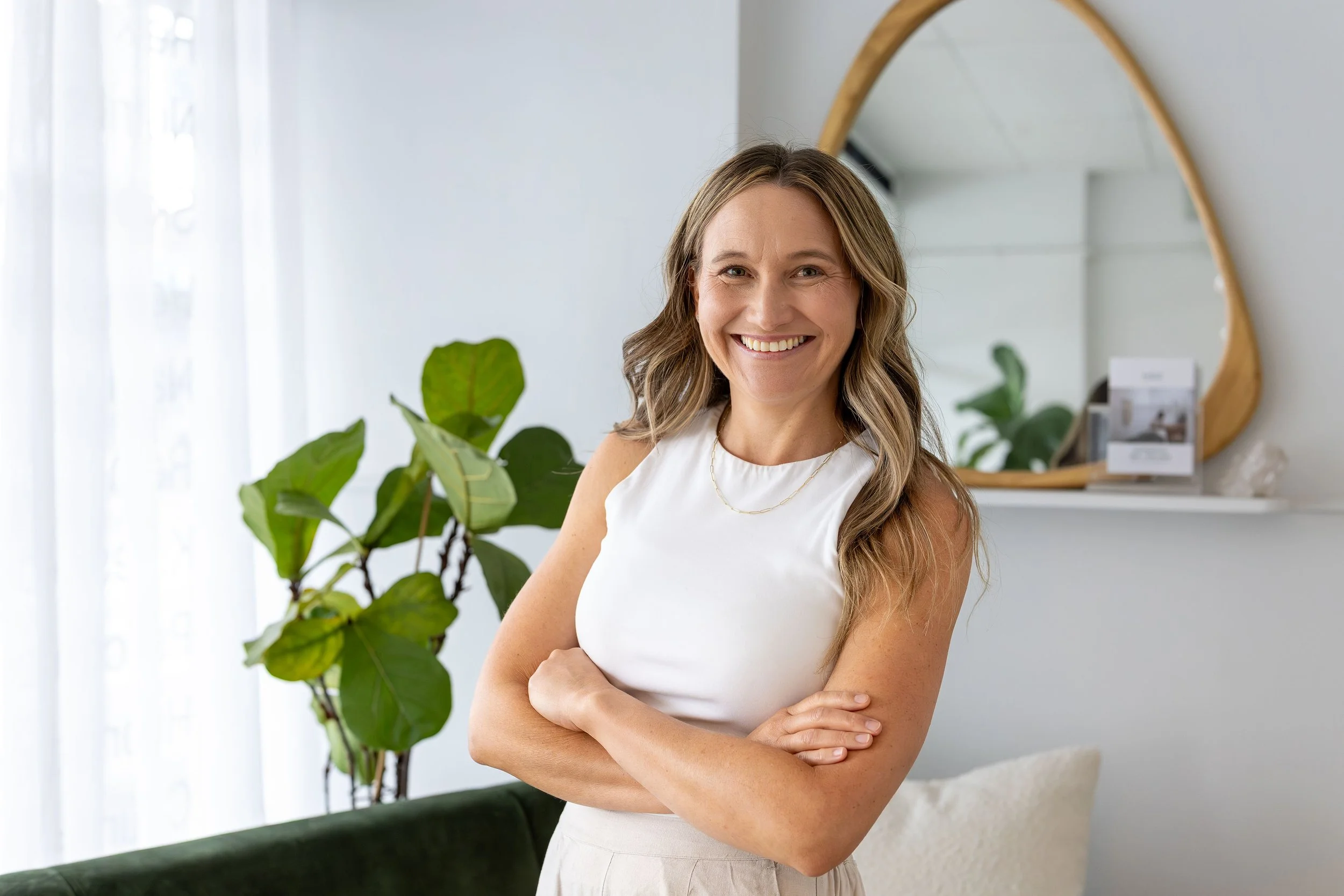 A smiling woman with long wavy hair wearing a white sleeveless top and beige pants, standing with crossed arms in a modern, well-lit room with large green plants and a mirror on the wall.