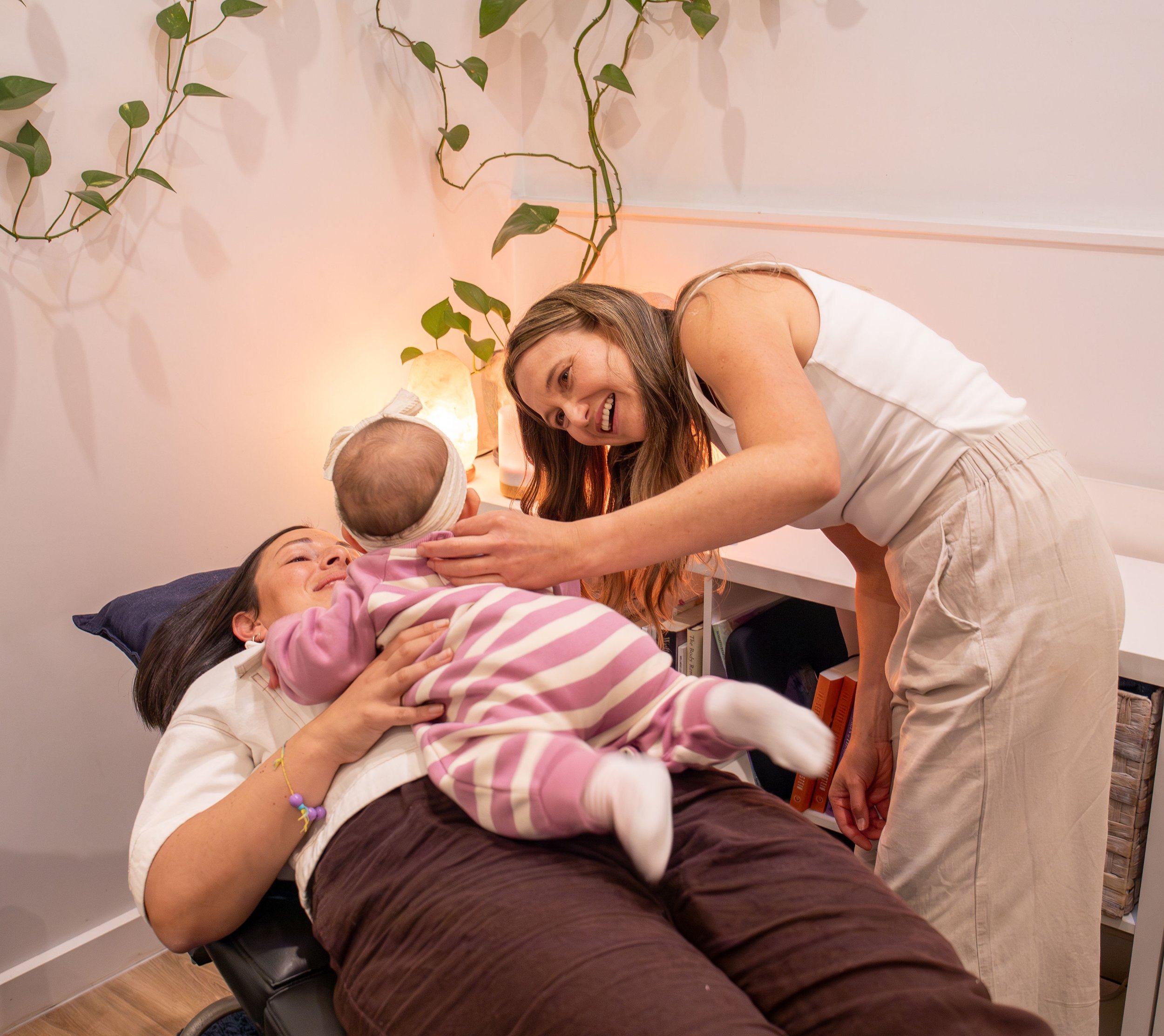 A woman with long dark hair holding a baby, lying on a bed with a woman with brown hair sitting beside them. They are indoors, with a pink wall, plant, and bookshelf in the background.
