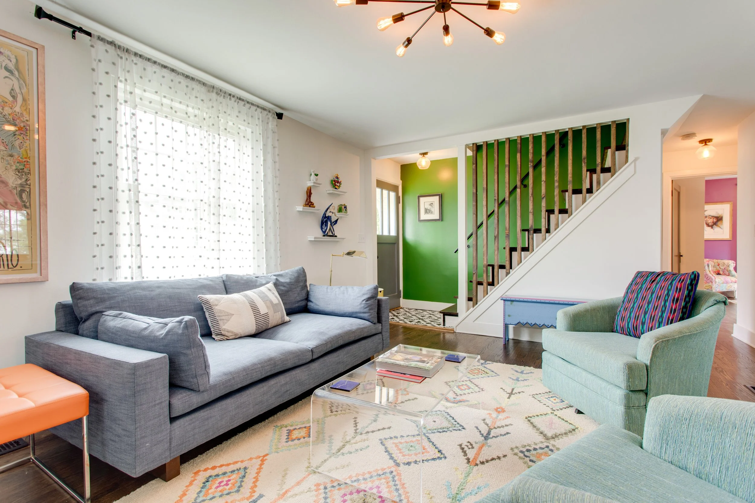 A living room featuring a gray sofa with pillows, a green armchair with a colorful striped pillow, a patterned rug, and a white coffee table with magazines. The room has large windows with sheer white curtains, a green accent wall with framed artwork, and a staircase with wooden slats. There are decorative shelves with figurines and a standing lamp near the sofa. A modern chandelier hangs from the ceiling.