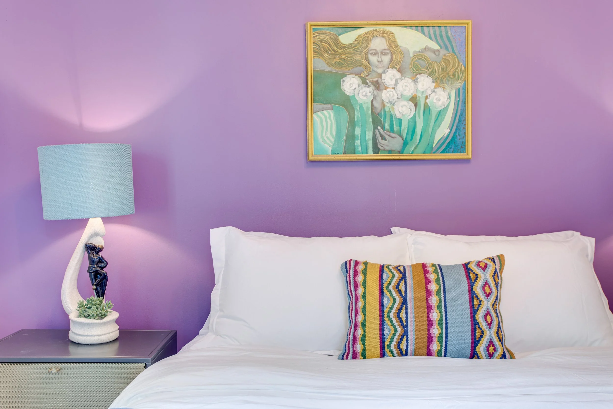 A bedroom with a purple wall, a white bed with a colorful embroidered pillow, a nightstand with a decorative lamp and a small plant, and a framed painting of a woman with flowing hair holding a bouquet of white flowers.
