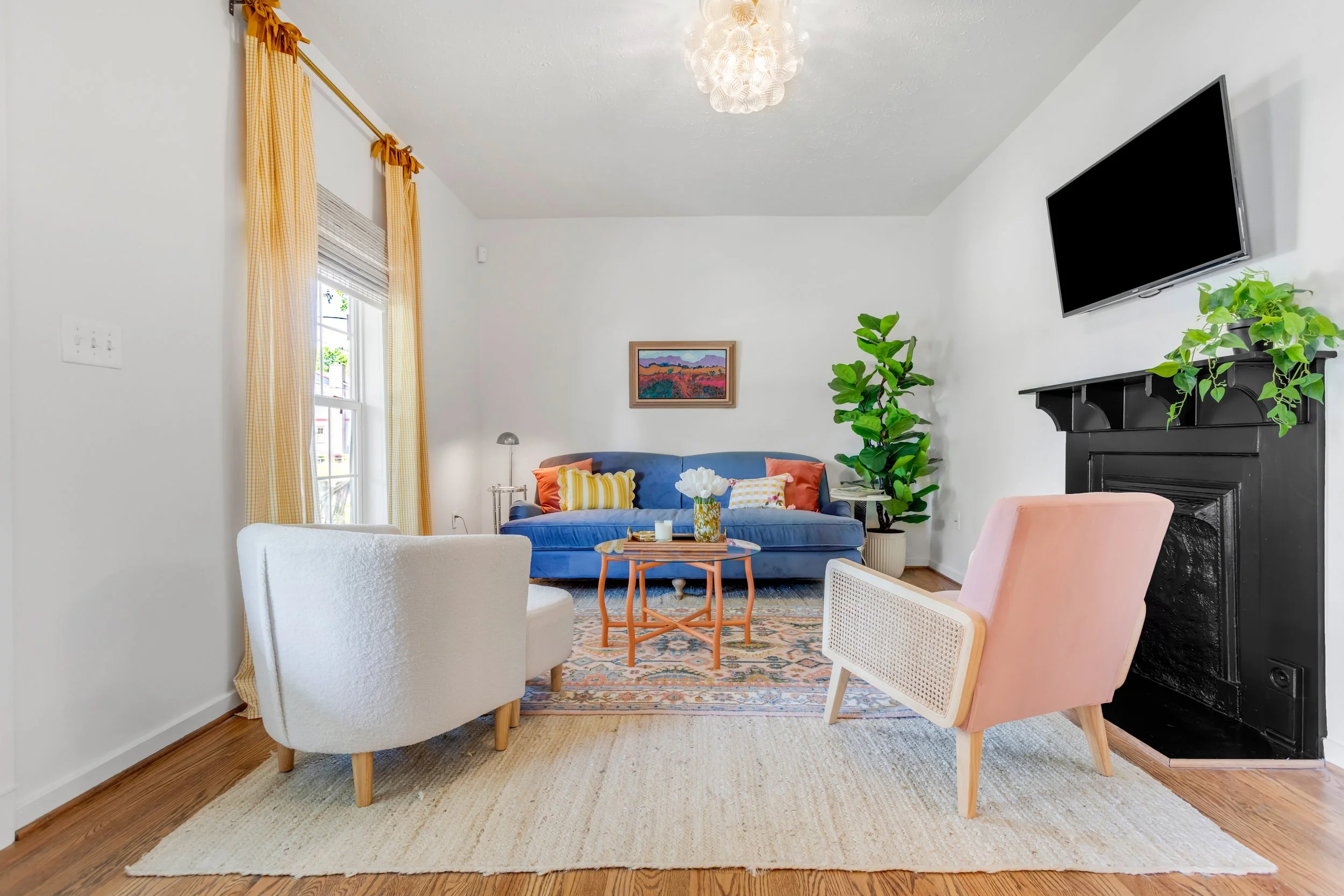 A living room with yellow curtains, a white armchair, a patterned area rug, a blue sofa with colorful pillows, a framed landscape painting, a green potted plant, a black fireplace, a wall-mounted TV, and a pink chair.