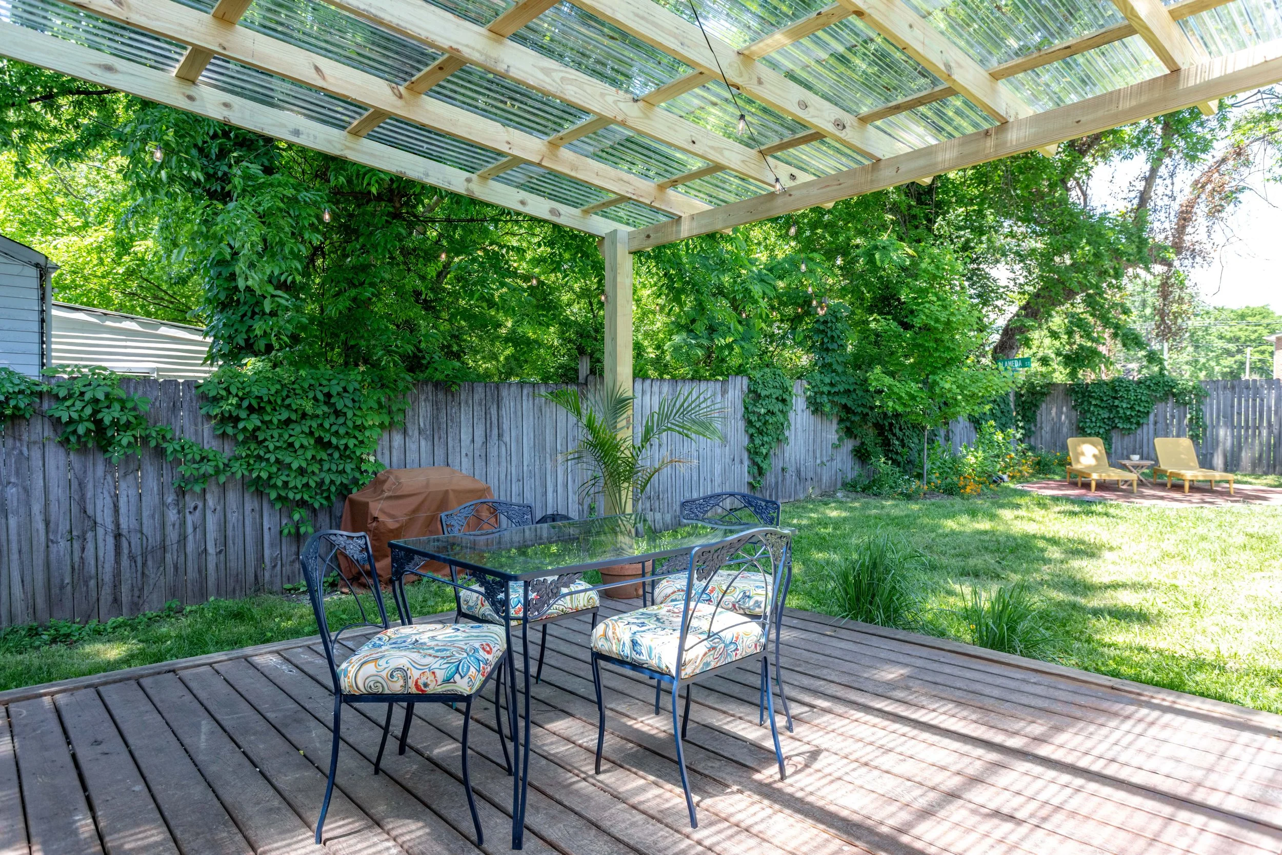 A backyard patio with a wooden deck, surrounded by green trees and plants, featuring a metal dining table with floral cushioned chairs, a small table with two chairs in the background, and a wooden fence with some climbing plants. There is a partiall