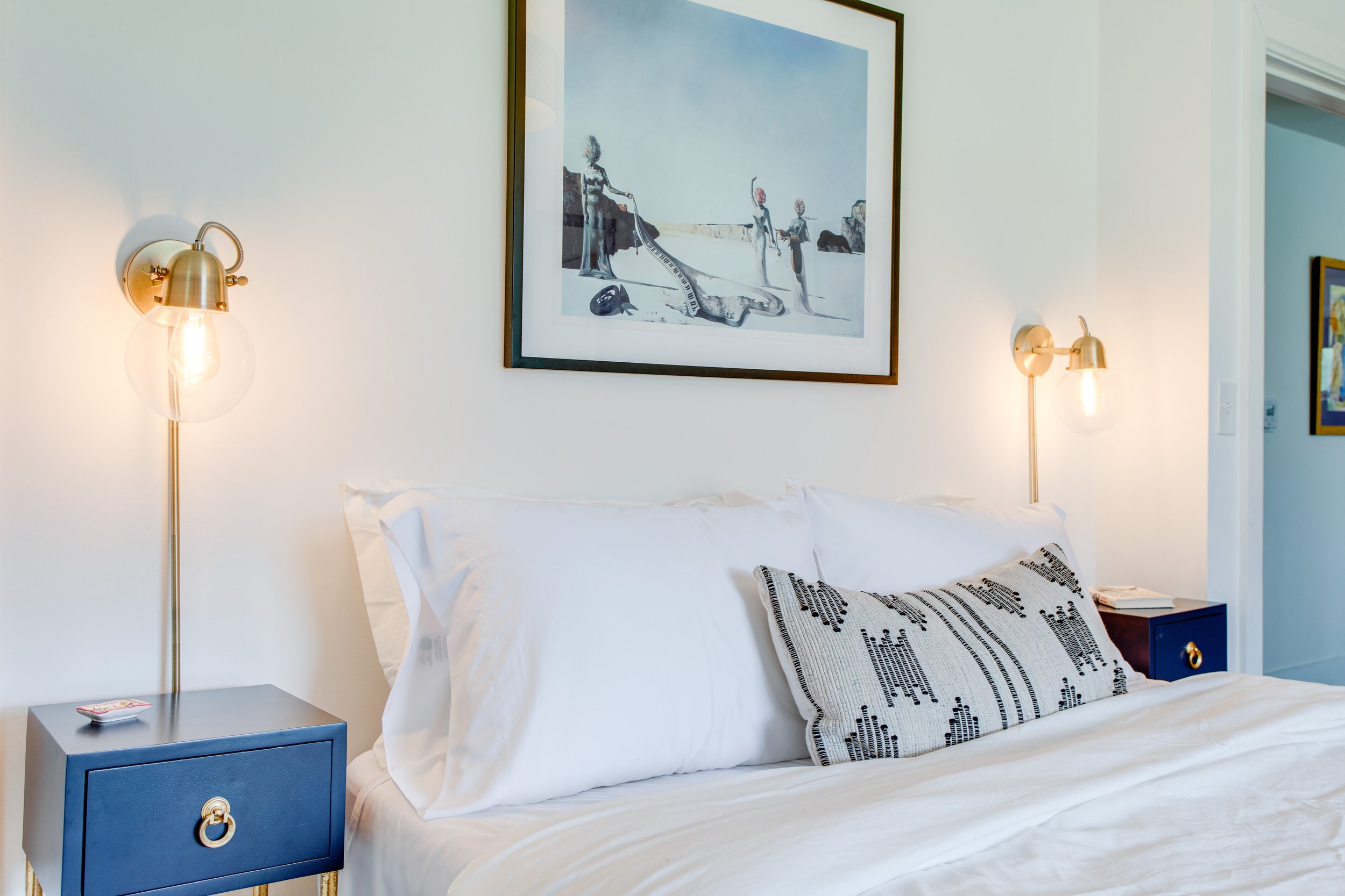 A bedroom with a white bed and decorative pillow, blue nightstand with a gold ring handle, wall-mounted gold lamps on each side of the bed, framed artwork of people at the beach above the bed, and a small book on the nightstand.
