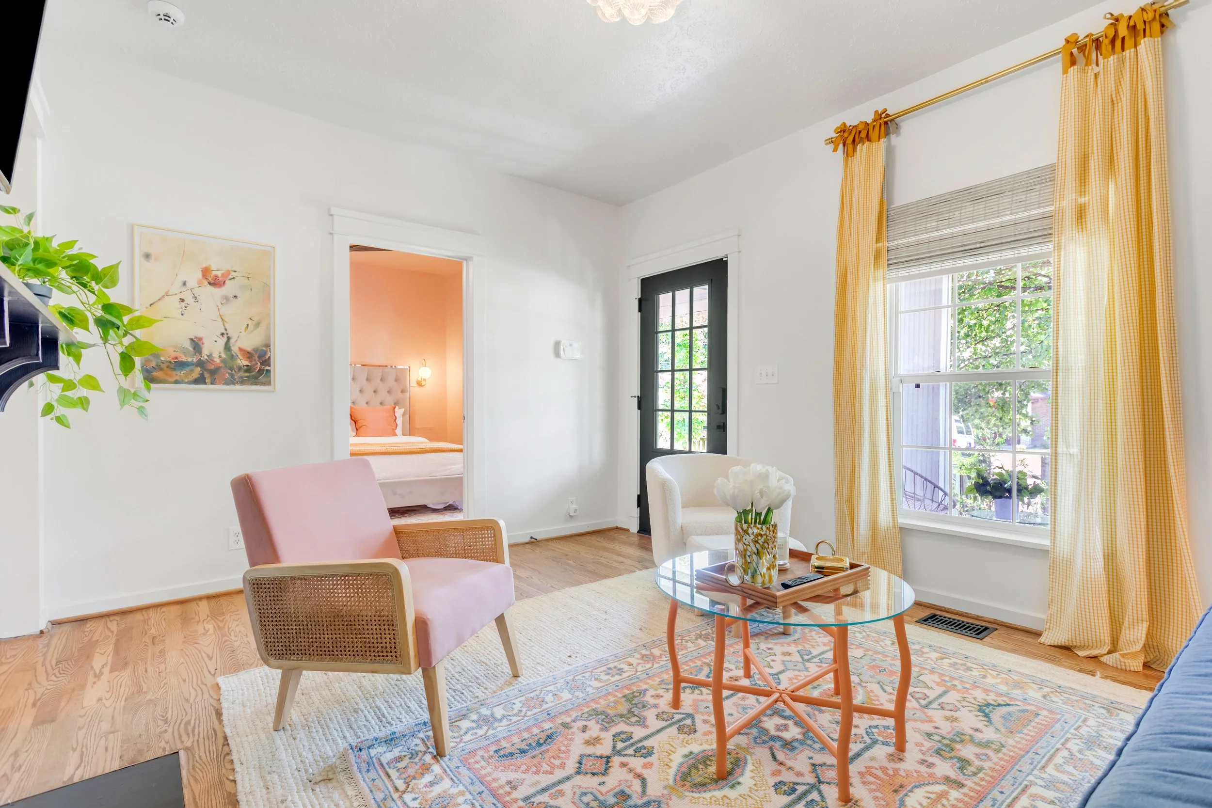 A cozy living room with white walls, a large window with yellow checkered curtains, and a patterned area rug. There is a pink armchair, a white armchair with a bouquet of flowers on a glass coffee table, and a small table with a tray. A doorway leads
