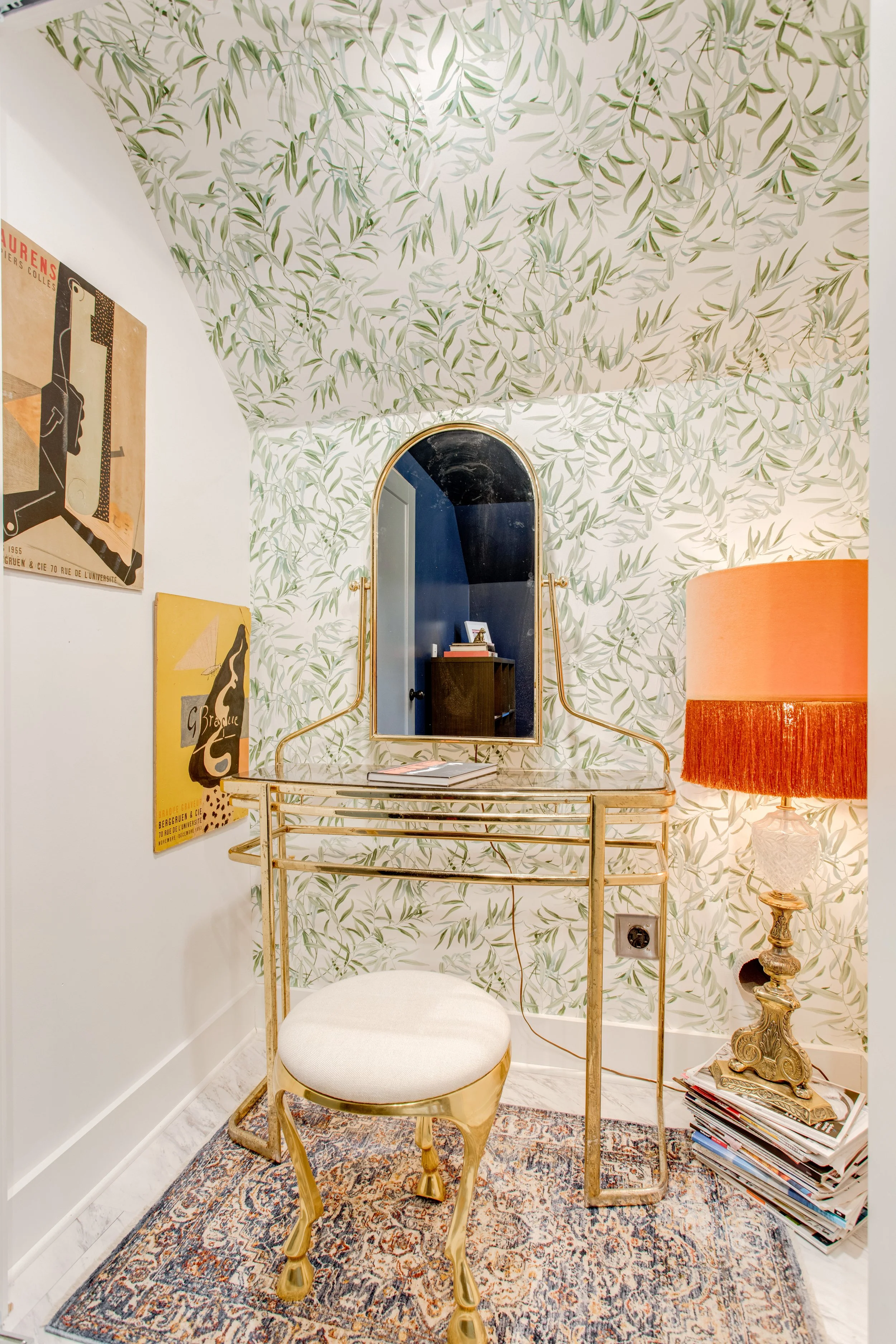 Decorative interior space with floral wallpaper, a gold-colored vanity table with a mirror, an upholstered stool, a table lamp with a large orange shade, and a stack of magazines on the floor nearby.