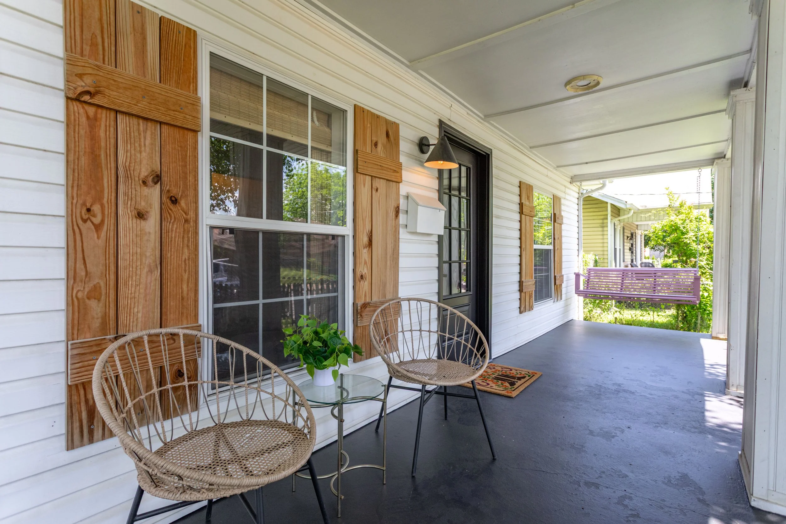 Porch with white siding, wooden shutters, black door, two wicker chairs, glass table with plant, purple bench, green trees in the background.