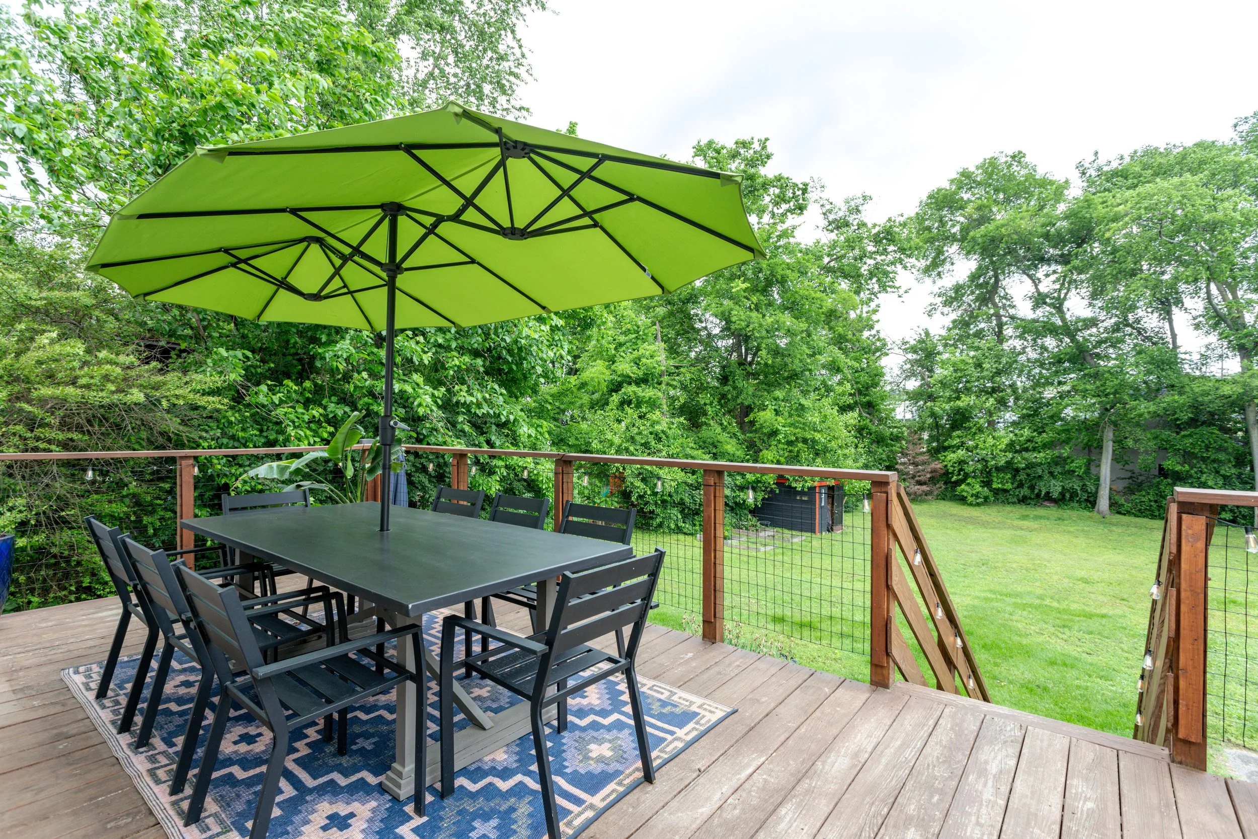 A wooden deck with a black outdoor dining table, six matching chairs, a green patio umbrella, on a patterned outdoor rug, surrounded by a wooden railing and lush green trees and grass in the background.