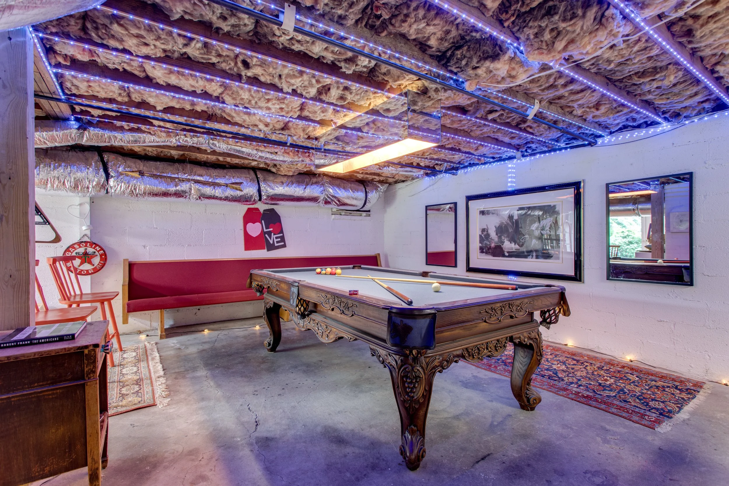 A basement game room with a decorative pool table, framed pictures, a red bench, and chairs. The ceiling is unfinished with exposed insulation and lighting, and the walls are painted white with various decorations.