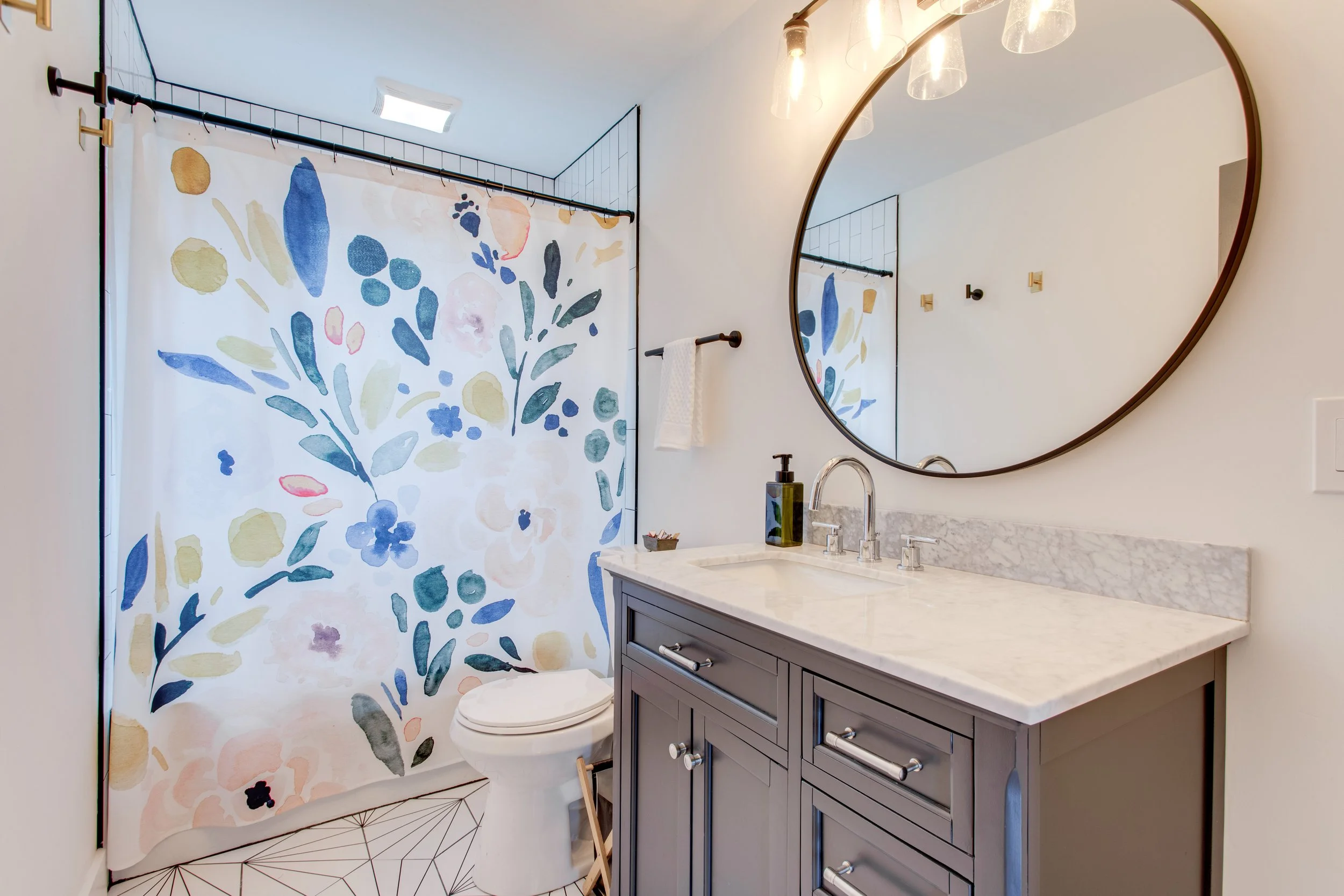 Bathroom with a round mirror, white walls, a gray vanity with silver knobs, a white marble countertop, a modern sink, a soap dispenser, and a shower with a colorful watercolor-style floral shower curtain. The floor has a geometric tile pattern.