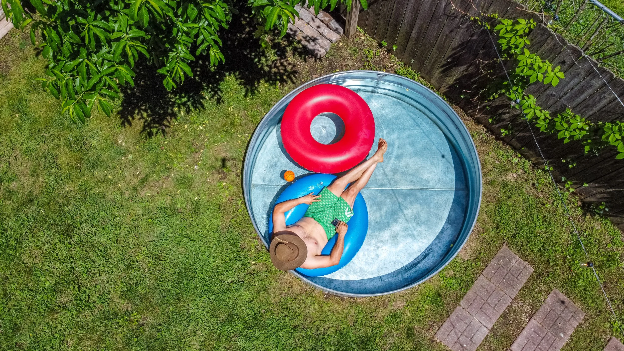 A person relaxing on a blue inflatable ring inside a small backyard pool with a red floating tube. The person is wearing green swim trunks, a brown hat, and sunglasses, while holding a smartphone. The pool is surrounded by grass and a wooden fence, w