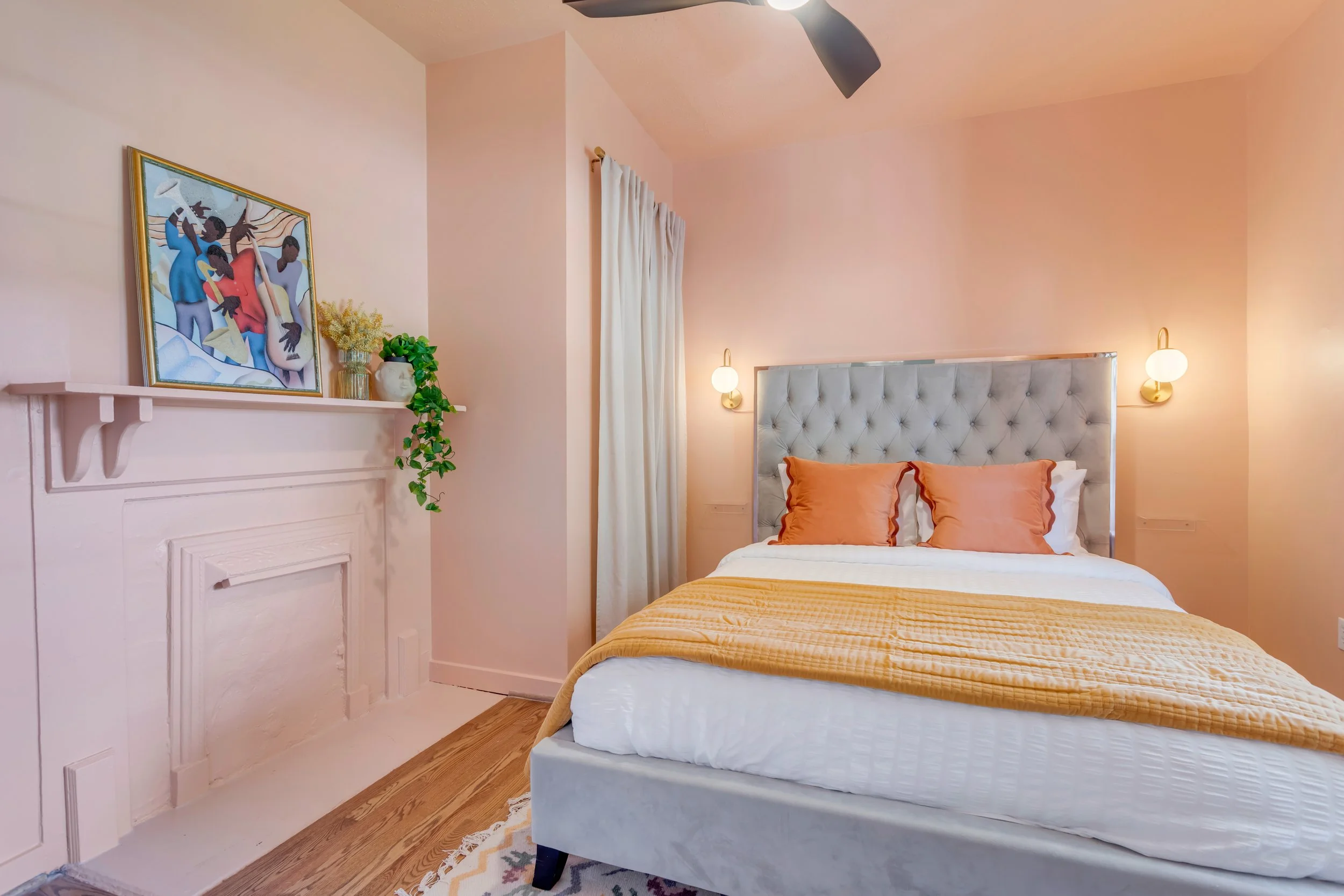 A bedroom with a pastel pink wall, a bed with white and orange bedding and a tufted headboard, two wall-mounted bedside lamps, a white curtain, a colorful abstract art piece, potted plants, and a hardwood floor.