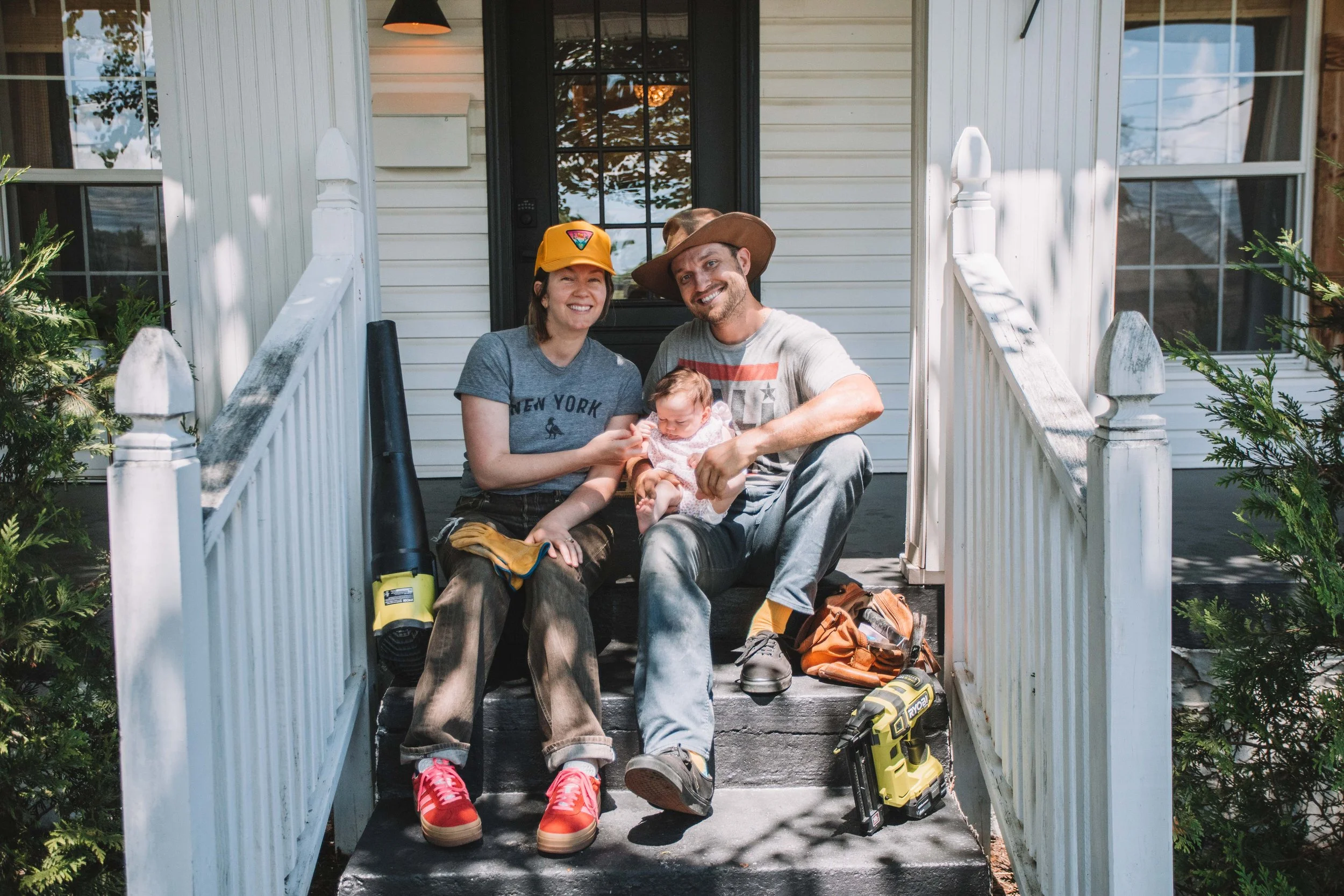 A family of three sitting on the front porch steps of their house, smiling. The woman wears a yellow cap and gray t-shirt, the man is wearing a cowboy hat and gray t-shirt, and their baby girl is in his lap. There are gardening tools and a backpack on the ground nearby.