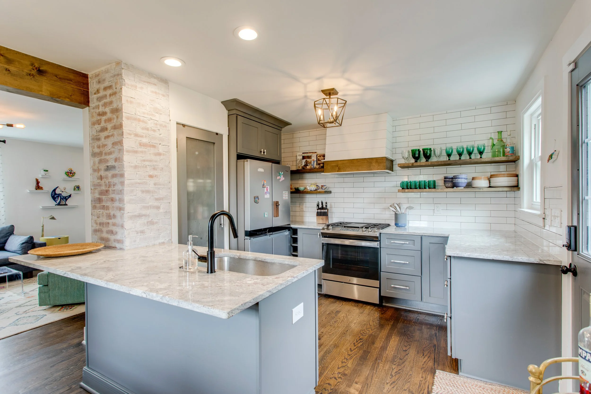 Modern kitchen with gray cabinetry, white subway tile backsplash, open wooden shelves with green and blue glassware, stainless steel appliances, a white marble countertop island, and a pendant light fixture.