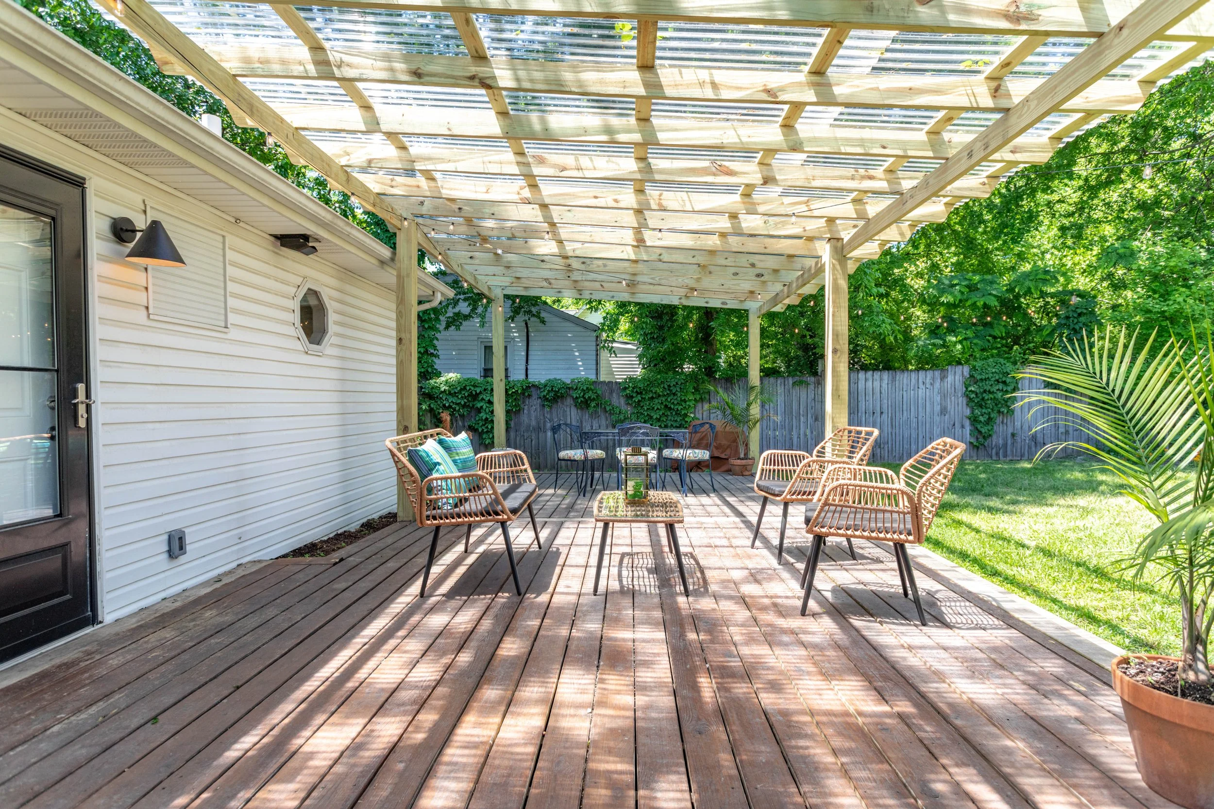 A backyard patio with a wooden deck and a pergola-style cover made of wooden beams with a transparent roof. There are outdoor chairs with cushions and a small table. The yard is surrounded by a wooden fence and lush green trees and plants.
