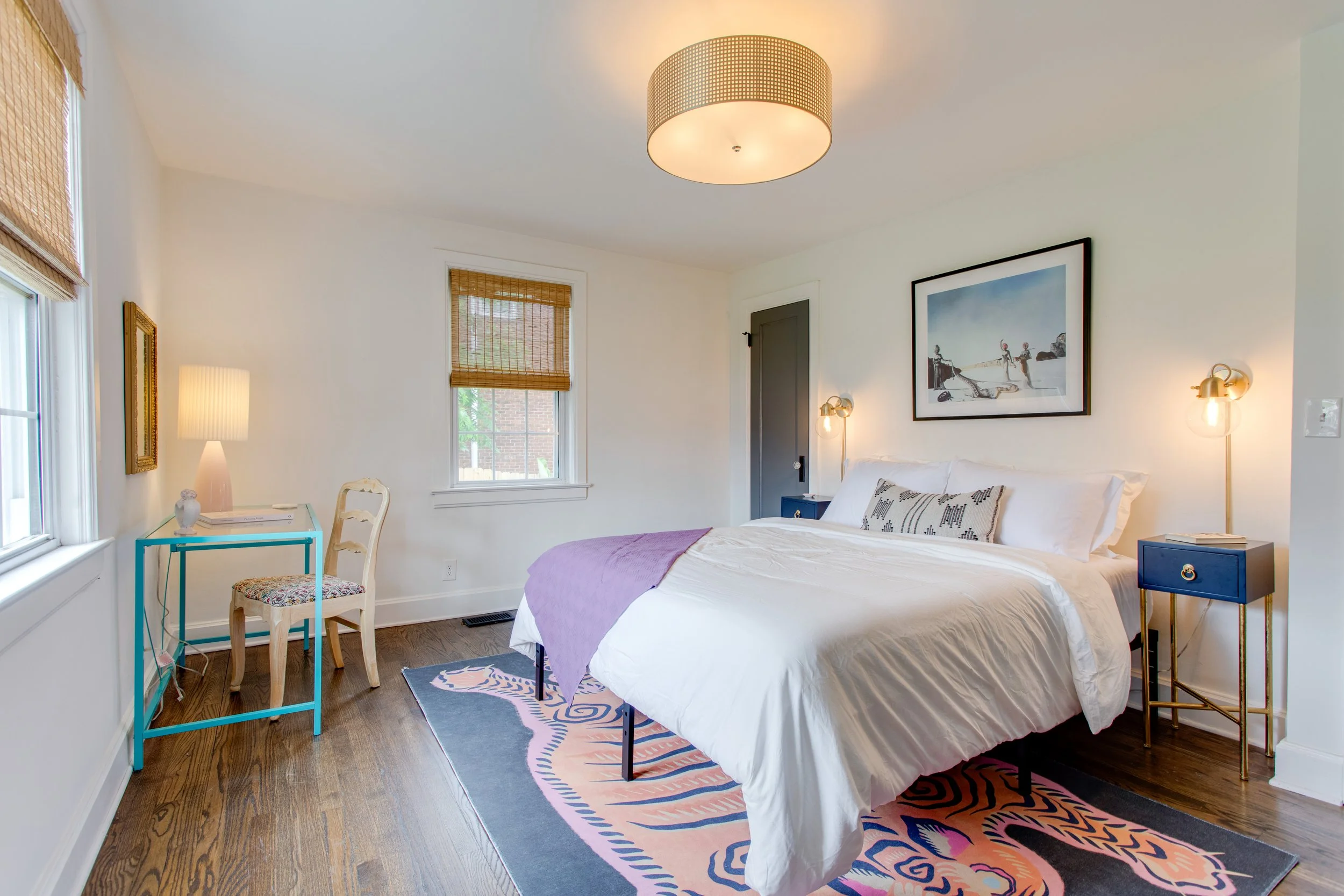 A bedroom with white walls, a large bed with white bedding, purple blanket, and patterned pillows. There are two windows with bamboo shades, a framed artwork above the bed, and a modern ceiling light fixture. Nightstand on the right side of the bed h