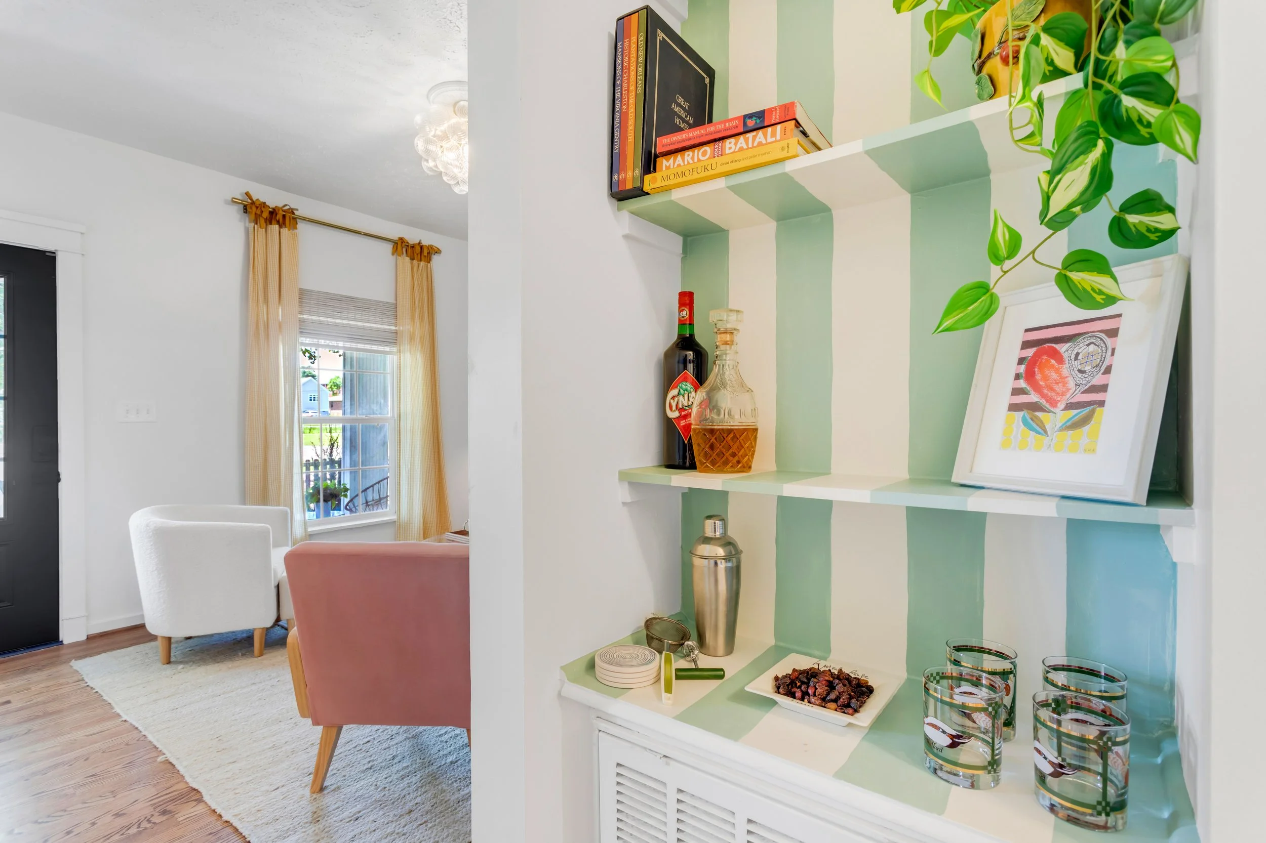 Interior of a living room with a window, yellow curtains, and a white armchair. A built-in corner shelf painted in green and white holds books, bottles, and a framed artwork, with some glassware and snacks on the lower shelf.