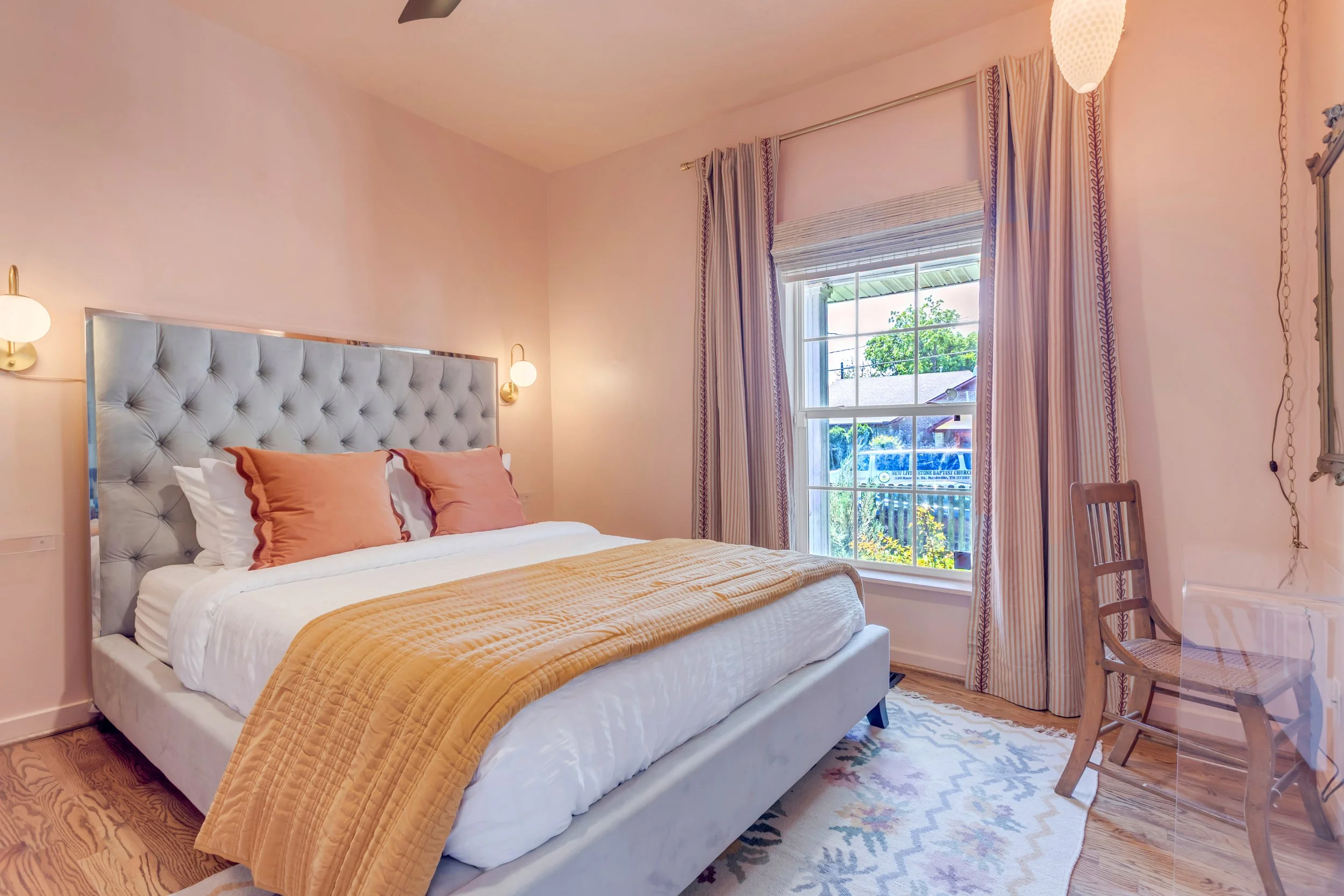 A bedroom with a bed that has a tufted headboard, white linens, and orange pillows. There are two wall-mounted round lamps on either side of the bed. A window with striped curtains and a view of trees and neighboring houses. A wooden chair is next to a glass table on the right.