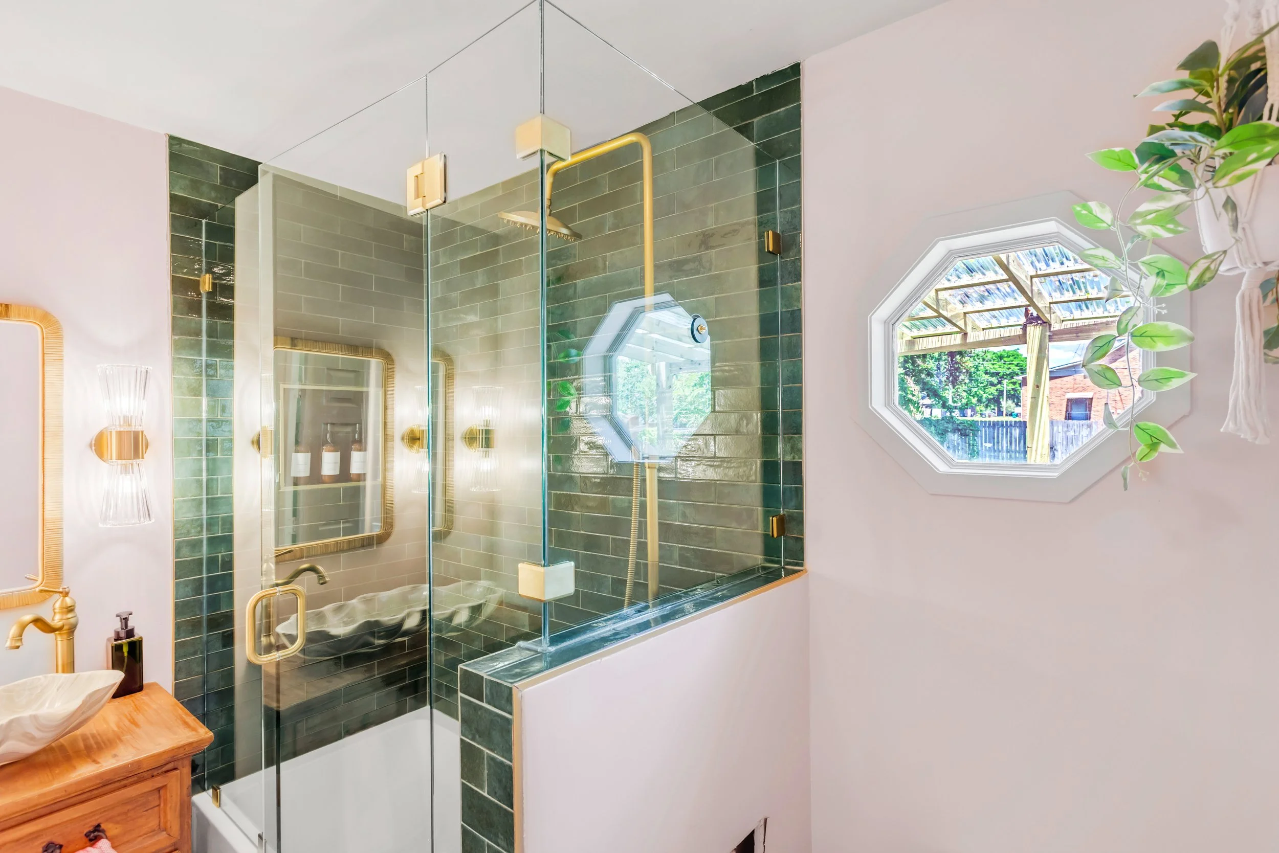 A bathroom with green tiles and a window showing a backyard scene, a glass shower enclosure with a rainfall showerhead, a wooden vanity with a vessel sink, and a plant near the window.