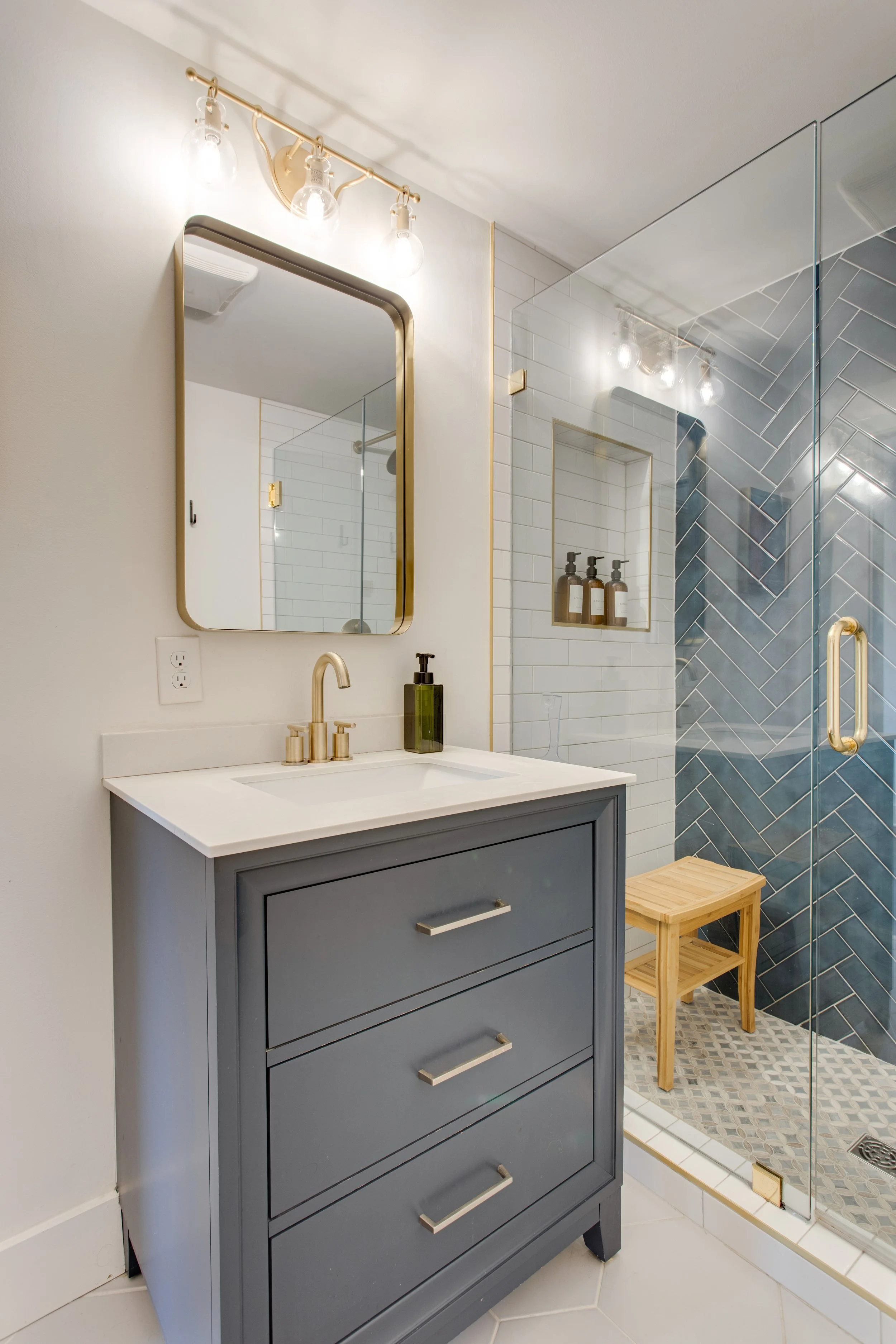 Bathroom with blue vanity, mirror, and mirrored shower enclosure with built-in shelf holding bottles, and a small wooden stool.