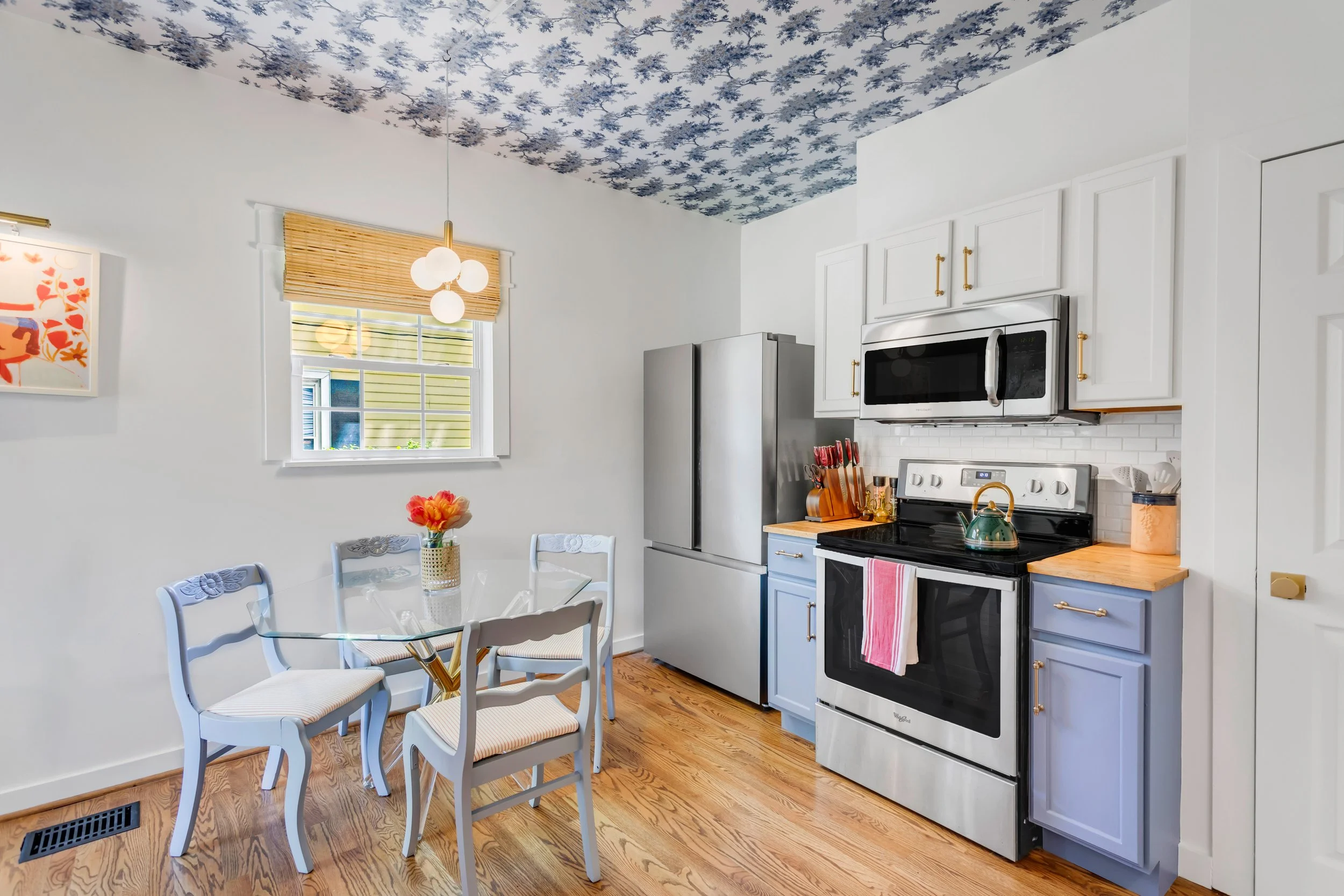 A bright kitchen with white walls and a blue ceiling featuring a floral pattern. There is a window with a bamboo blind and a modern chandelier hanging nearby. The kitchen has a white stove, a stainless steel refrigerator, and white cabinets with gold