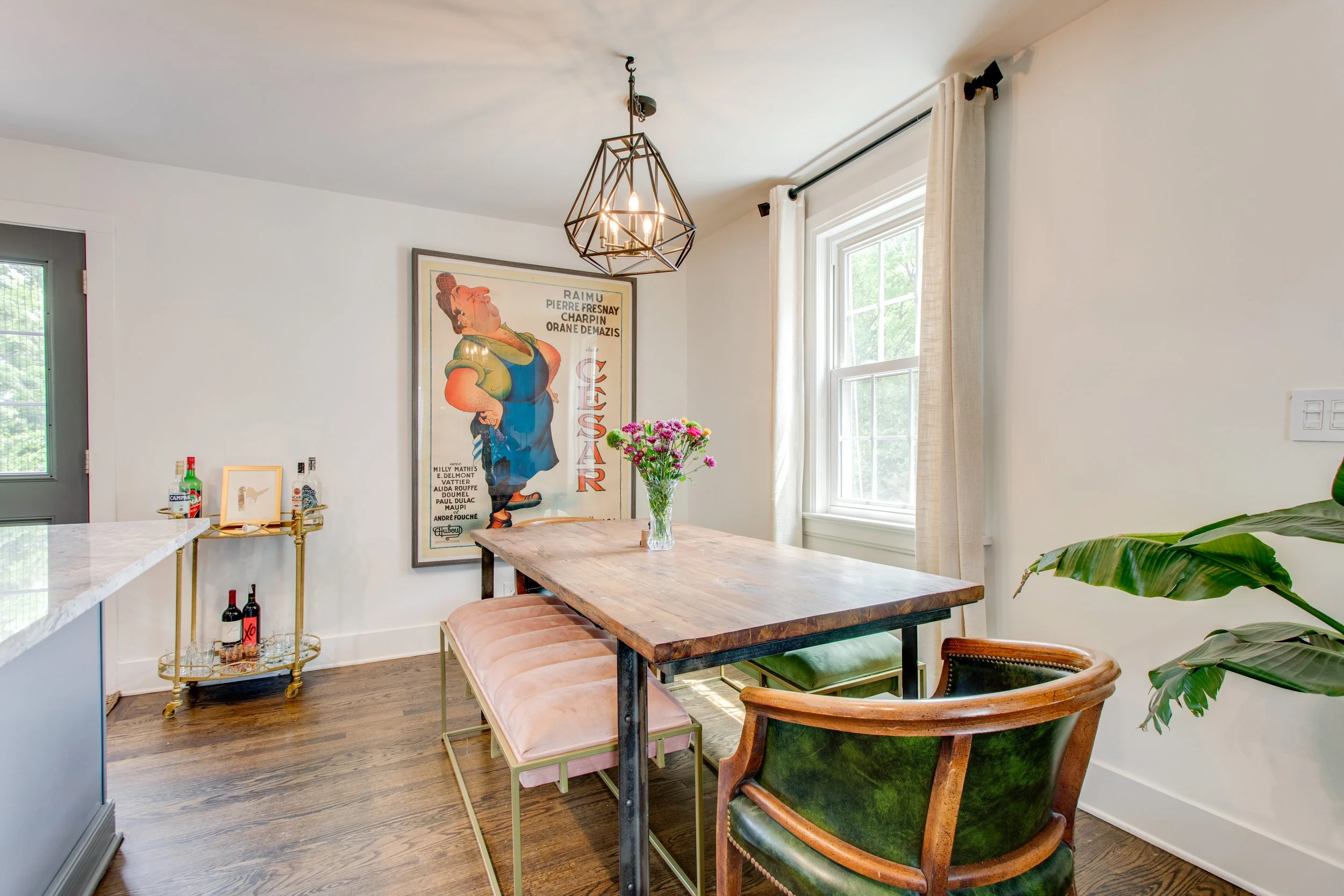 A dining room with a wooden table, pink velvet bench, and green velvet armchair, large window with white curtains, a framed poster on the wall, a glass vase with colorful flowers on the table, and a geometric metal chandelier hanging from the ceiling
