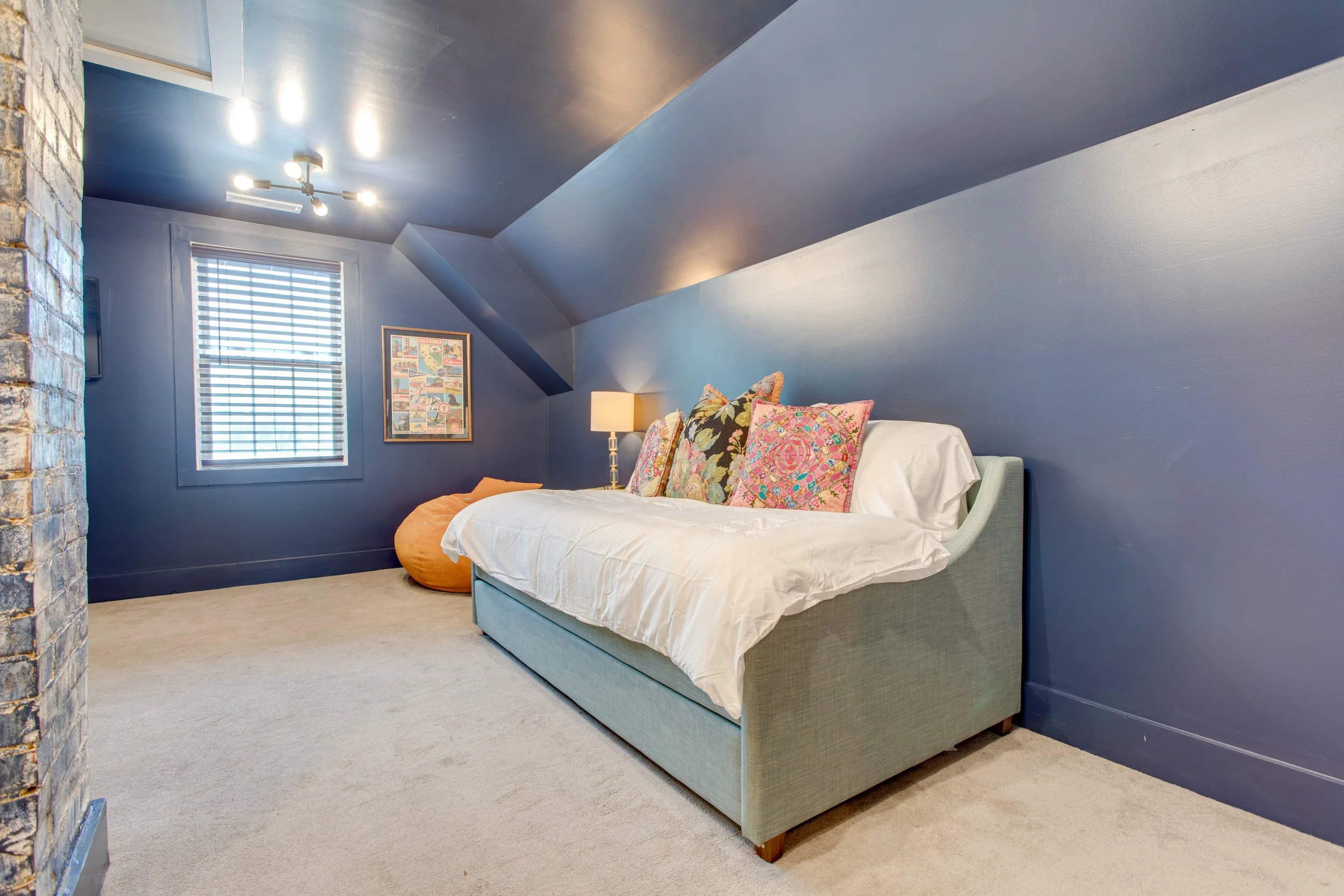 A bedroom with blue walls and ceiling, featuring a bed with a white cover, colorful decorative pillows, a wooden nightstand with a lamp, a framed art piece, a window with blinds, and a brick wall on the left side.