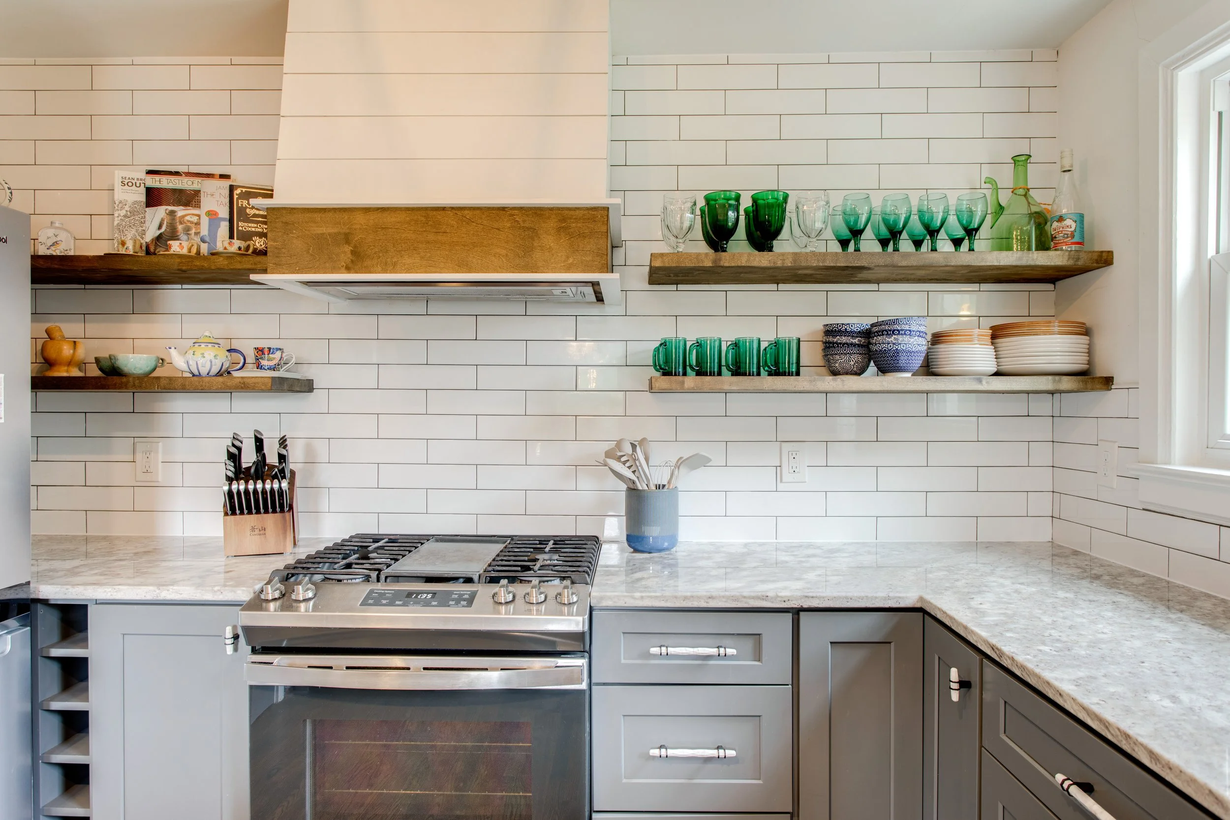 A modern kitchen with white subway tile backsplash, gray cabinets, and a marble countertop. There are wooden open shelves with various dishes, glasses, and cookbooks. A stainless steel stove is integrated into the counter, and a blue ceramic container holds kitchen utensils. Natural light comes from a window on the right.