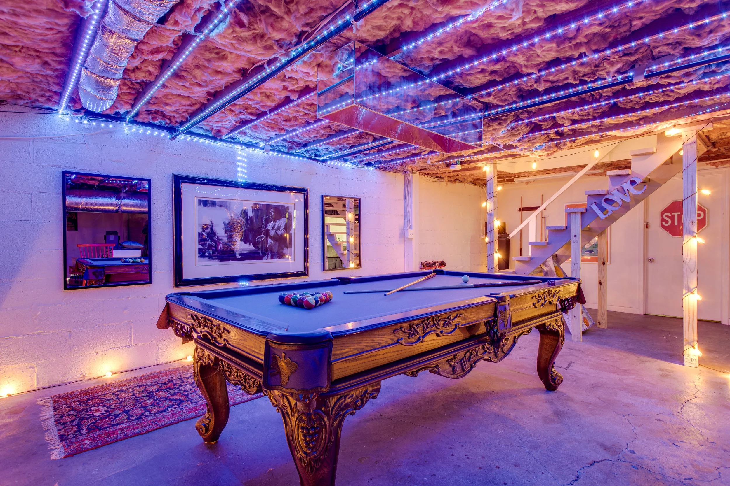 A basement with exposed pink insulation on the ceiling, string lights, a pool table with ornate wood carvings, a large mirror, and a staircase with a 