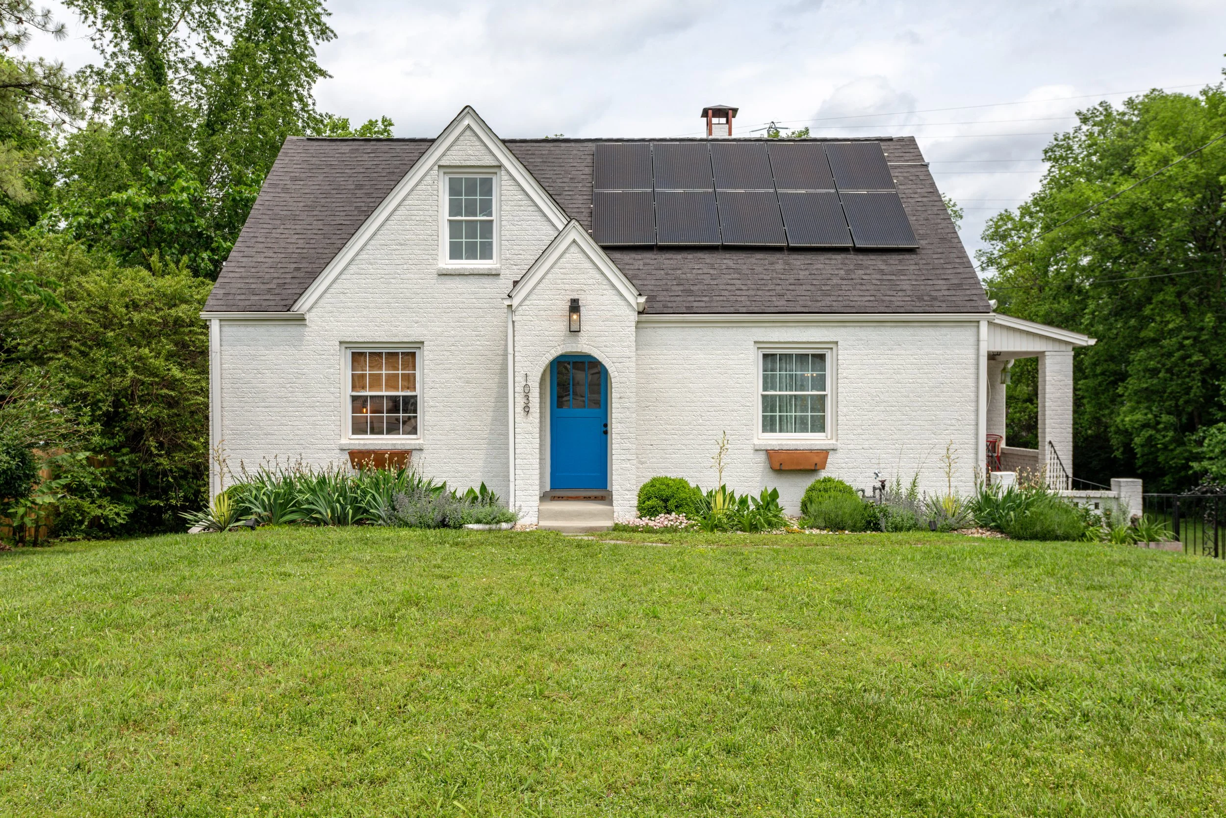 Cozy white home with blue front door and solar panels on the roof, surrounded by a green lawn, bespoke year-round garden and huge oak & pine trees.