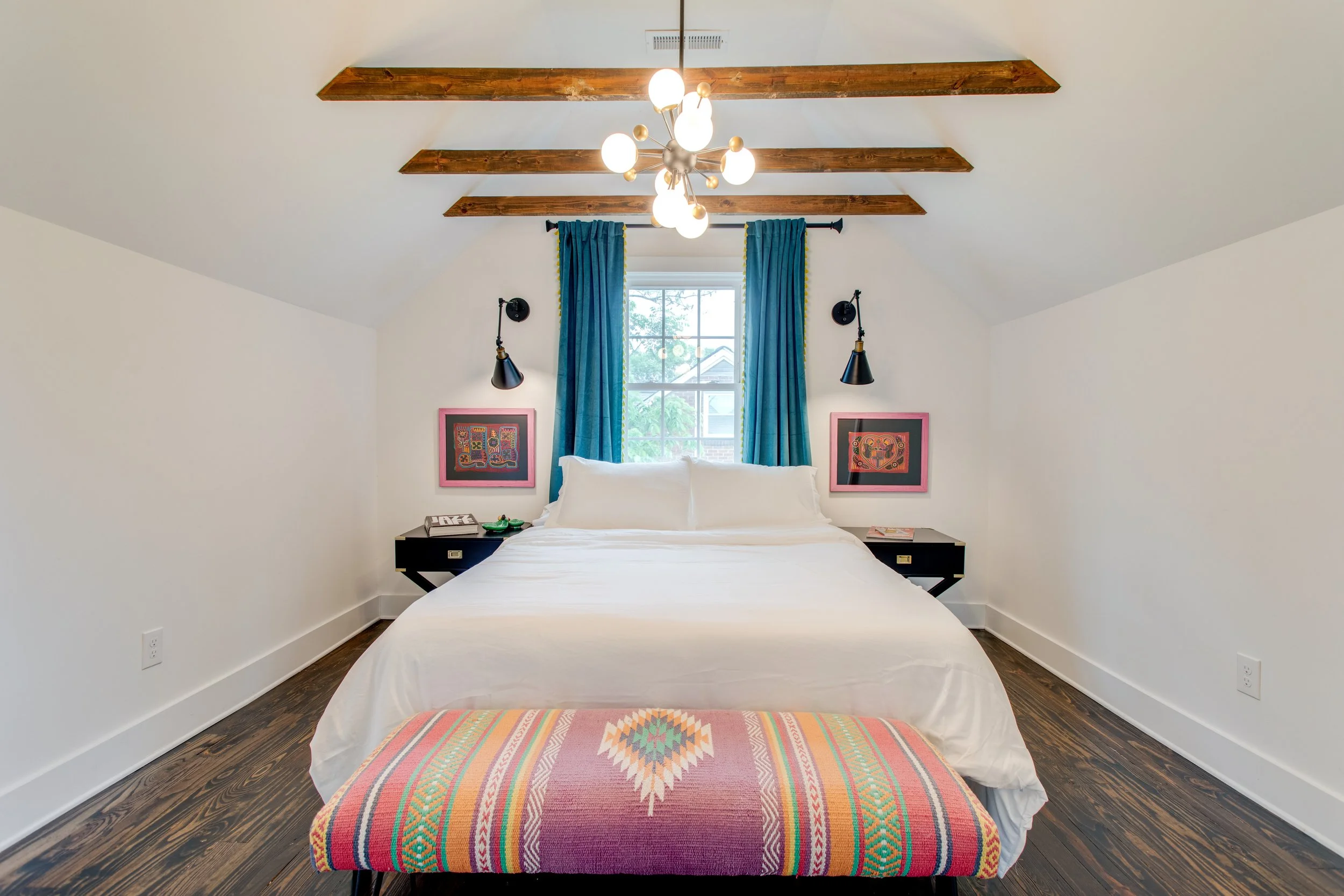 Bedroom with white walls, a bed with white linens, two black nightstands, and a colorful woven rug at the foot of the bed. Blue curtains hang on either side of a window with a view outside. A modern chandelier hangs from the ceiling with wooden beams