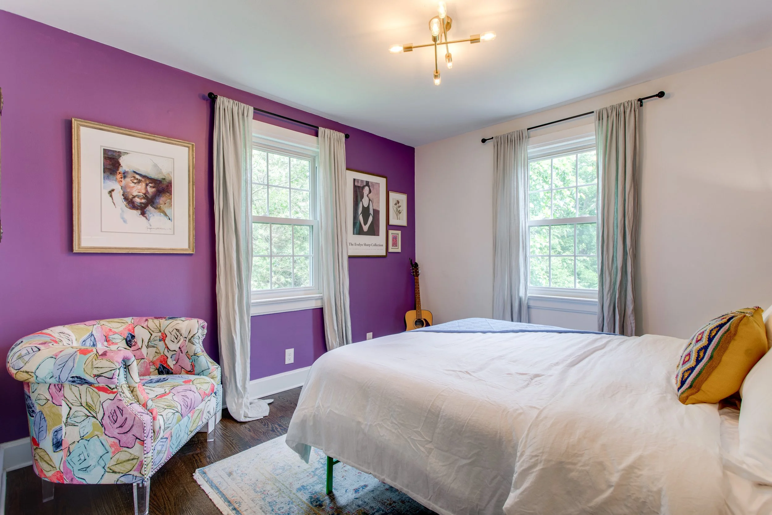 A bedroom with a purple accent wall, light-colored curtains, two windows, and framed artwork, including a portrait of a man and a woman, along with a guitar in the corner, a bed with white bedding, a colorful armchair, and a chandelier overhead.