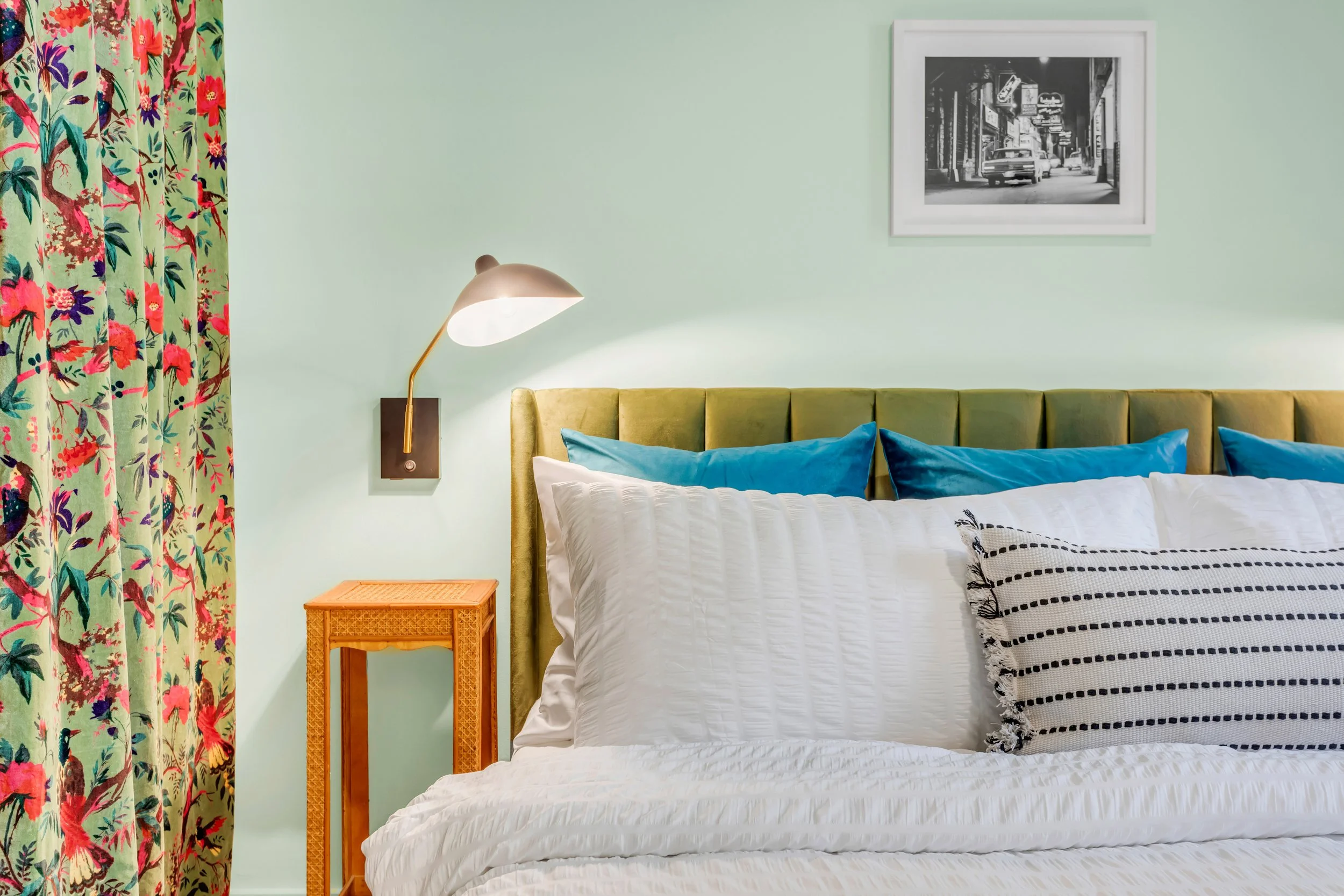 A bedroom with a green wall, a yellow upholstered headboard, white bedding, and black and white striped pillows. On the left, vibrant floral curtain with pink, red, purple, and green patterns. Next to the bed, a small wooden nightstand with a wicker 