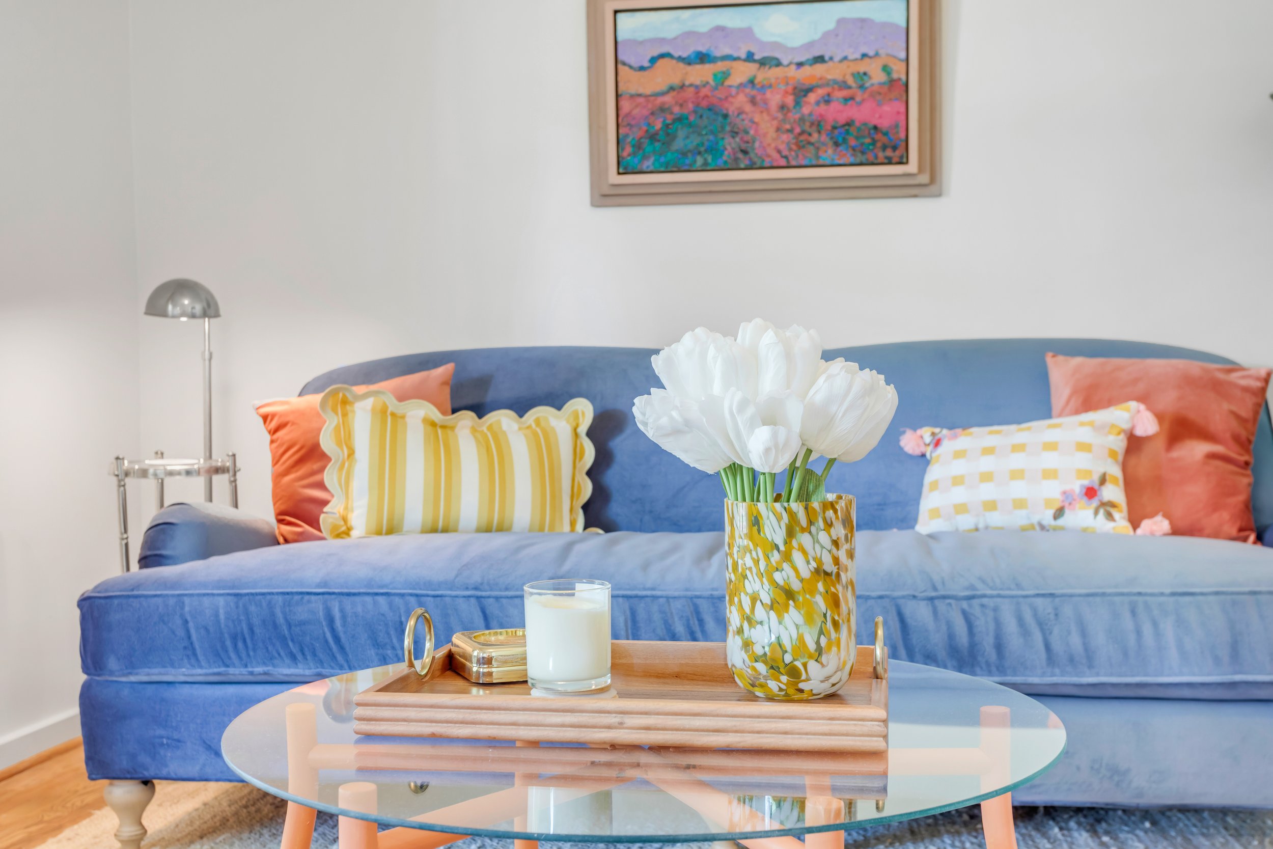 Living room scene with a blue velvet couch decorated with colorful pillows, a wooden coffee table with a tray, a candle, and a yellow flower vase with white flowers. A framed landscape painting hangs on the white wall above the couch.