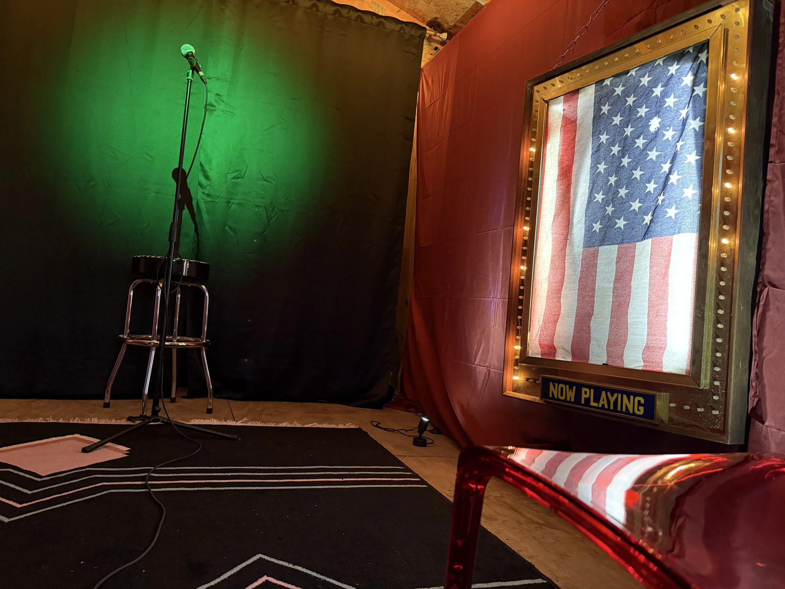 A stage with a microphone on a stand and a stool, a backdrop with green lighting, and a corner with a red curtain and a display case with an American flag, decorated with string lights and a sign that reads "NOW PLAYING".